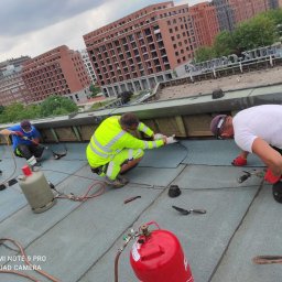 Artur Górak Backup-Dach - Trzech dekarzy w trakcie krycia papą termozgrzewalną płaskiego dachu budynku miejskiego, widoczne butle gazowe, narzędzia i budynki w tle.