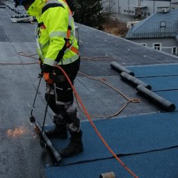 Artur Górak Backup-Dach - Pracownik w odblaskowej kurtce i kasku spawa papę na dachu płaskim, widoczny palnik, rolki papy i młotek dekarski, w tle budynki i drzewa.