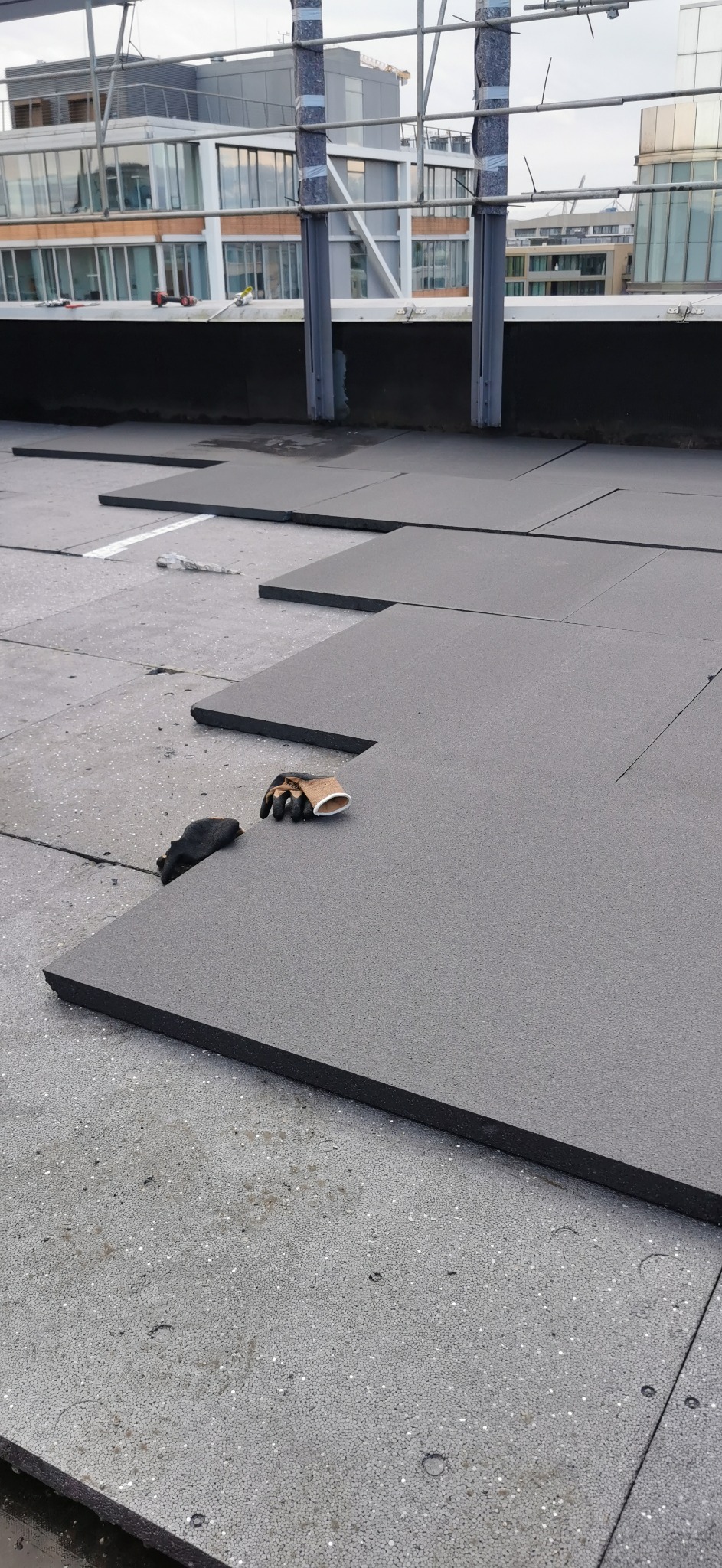 Układanie szarych płyt izolacyjnych na dachu, widoczne rękawice robocze obok jednej z płyt, w tle budynki miejskie i metalowa konstrukcja.