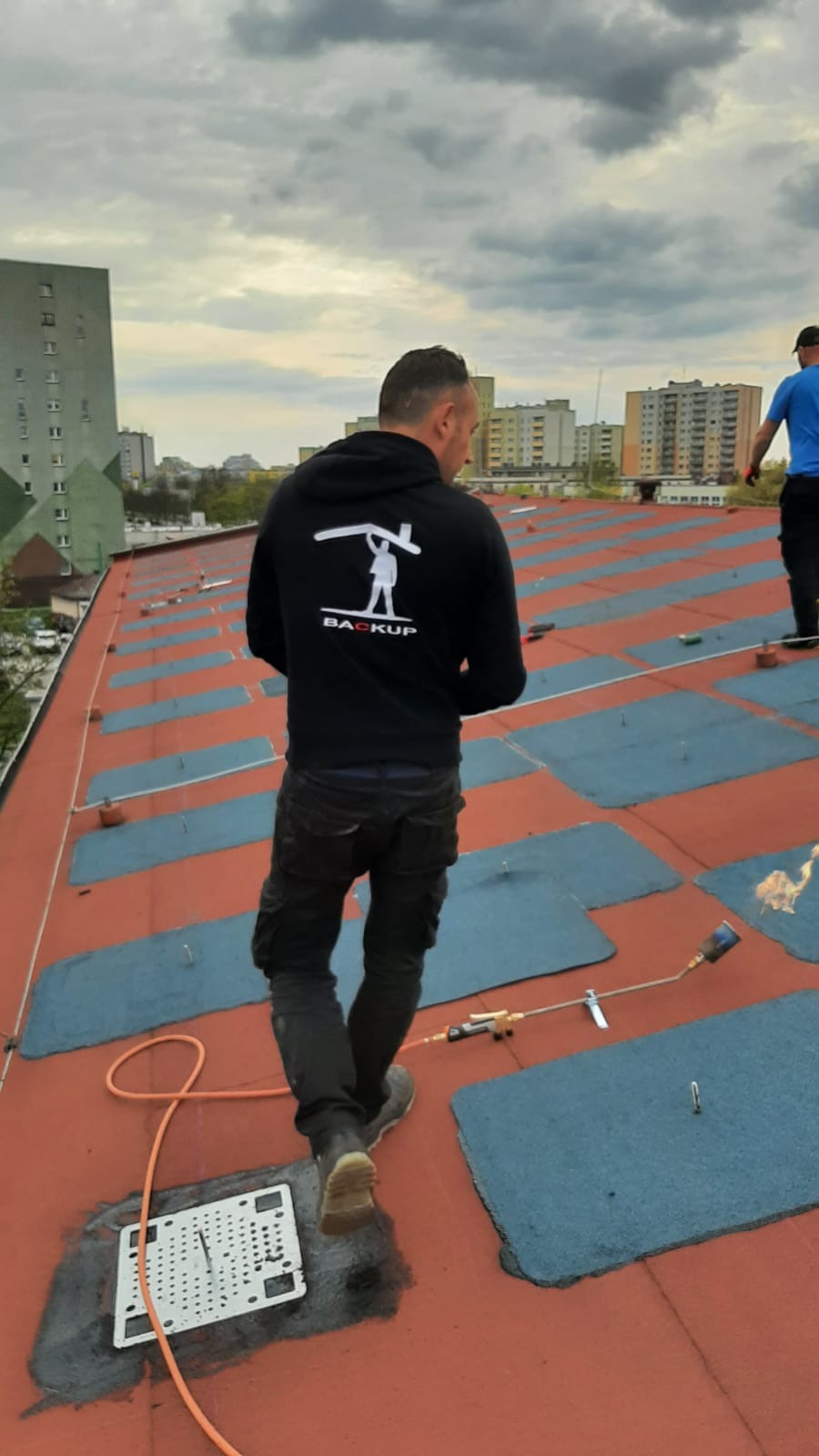 Pracownik w bluzie z logo firmy na dachu pokrytym papą termozgrzewalną, w tle budynki mieszkalne, widoczny palnik dekarski w trakcie pracy.