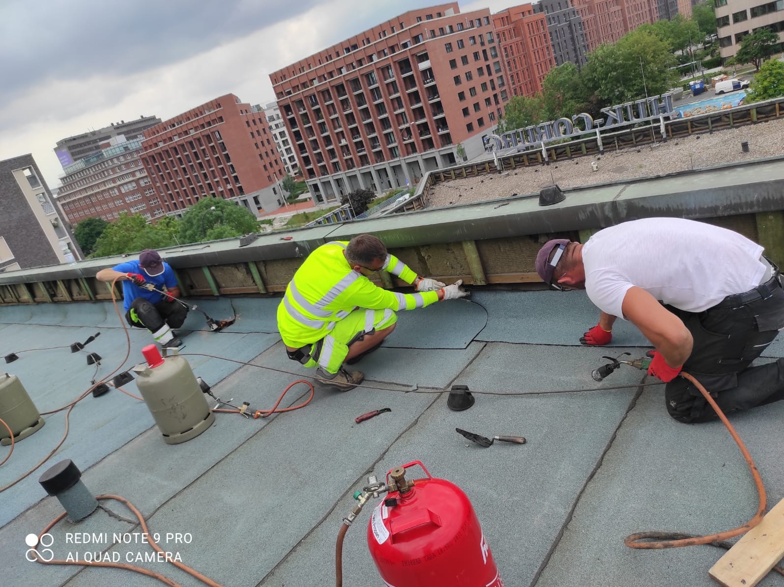 Trzech dekarzy w trakcie krycia papą termozgrzewalną płaskiego dachu budynku miejskiego, widoczne butle gazowe, narzędzia i budynki w tle.