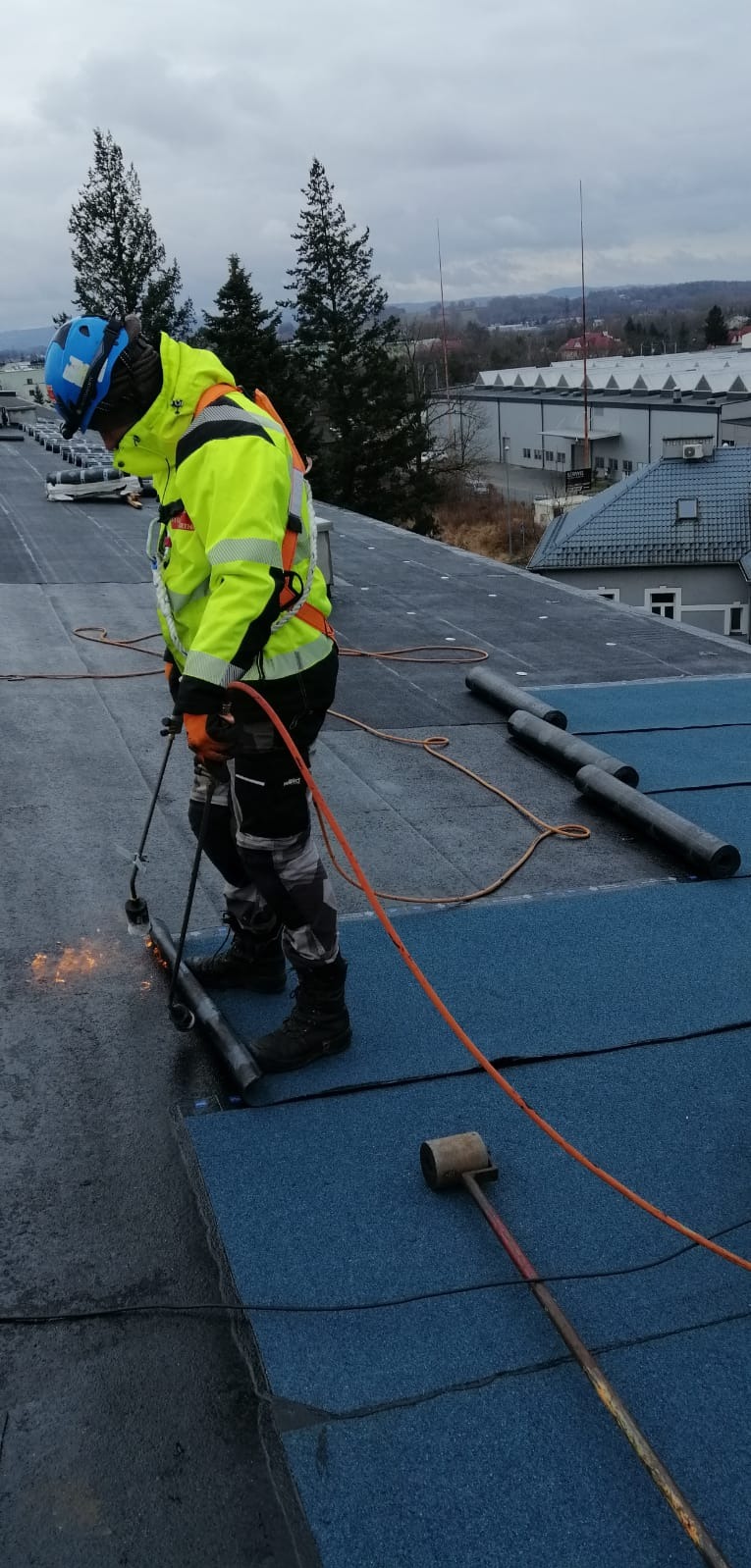 Pracownik w odblaskowej kurtce i kasku spawa papę na dachu płaskim, widoczny palnik, rolki papy i młotek dekarski, w tle budynki i drzewa.