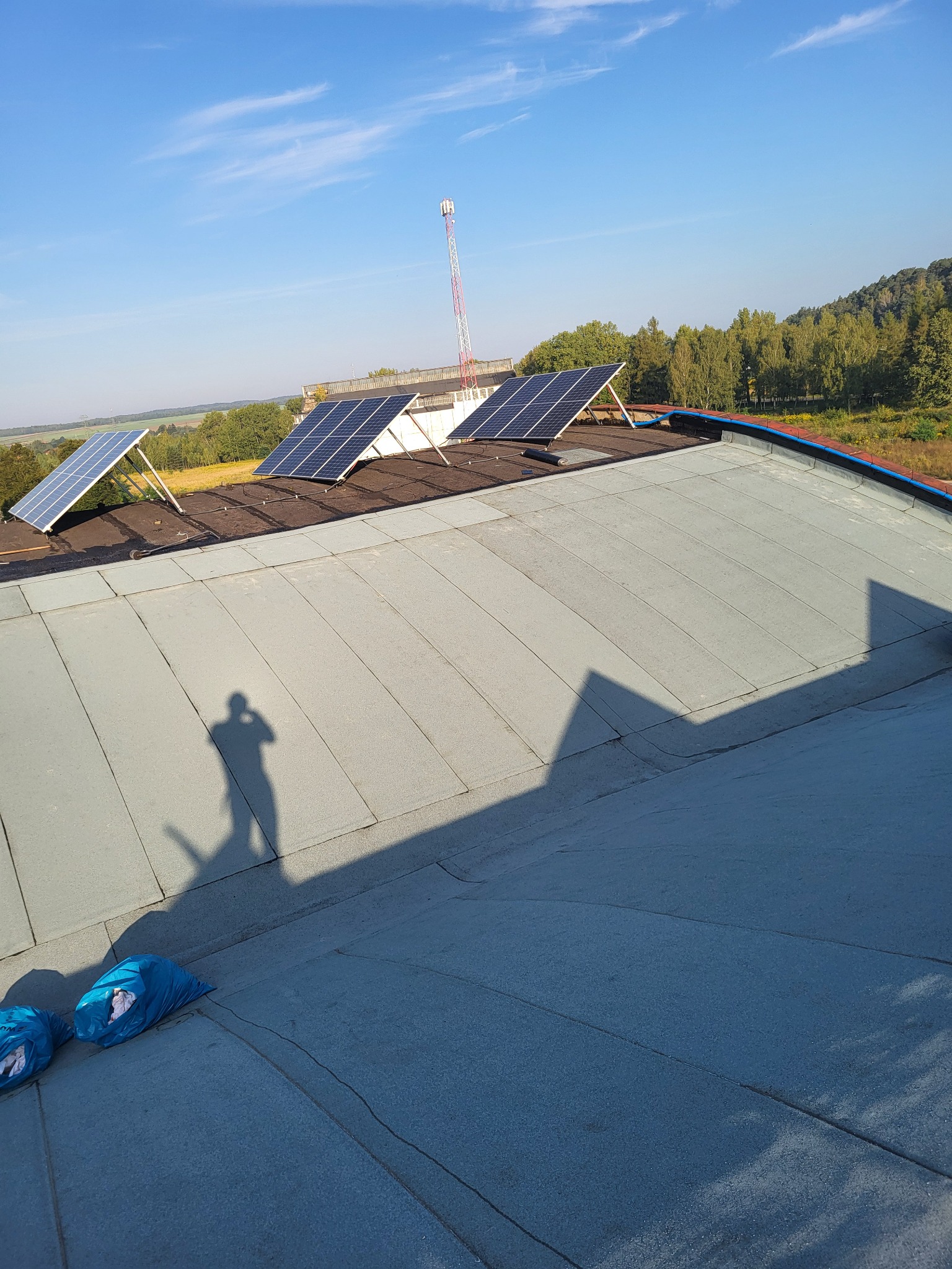 Płaski dach pokryty szarą papą z zamontowanymi panelami słonecznymi; w rogu widoczne worki z odpadami budowlanymi, cień fotografa.