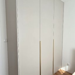 MACIEJ KLUK - Jasna, minimalistyczna szafa z czterema drzwiami i długimi, złotymi uchwytami, dopasowana do wnętrza sypialni z drewnianą podłogą i widocznym fragmentem łóżka.