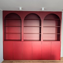 MACIEJ KLUK - Czerwona zabudowa meblowa z trzema przeszklonymi, łukowatymi wnękami i szafkami na dole, na tle drewnianej podłogi. Minimalistyczny design, nowoczesne oświetlenie.