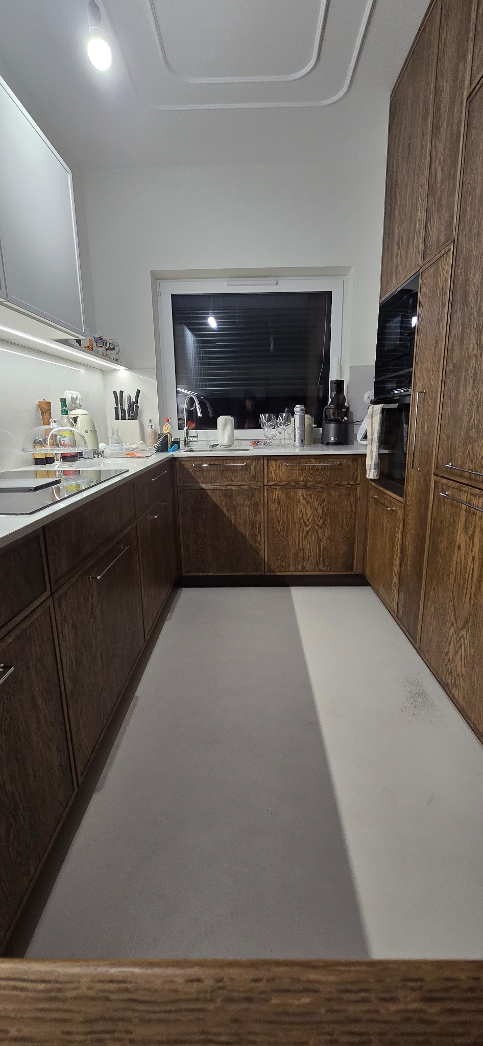 Wnętrze kuchni z drewnianymi szafkami, jasnym blatem i oknem z żaluzjami, oświetlone lampą sufitową.