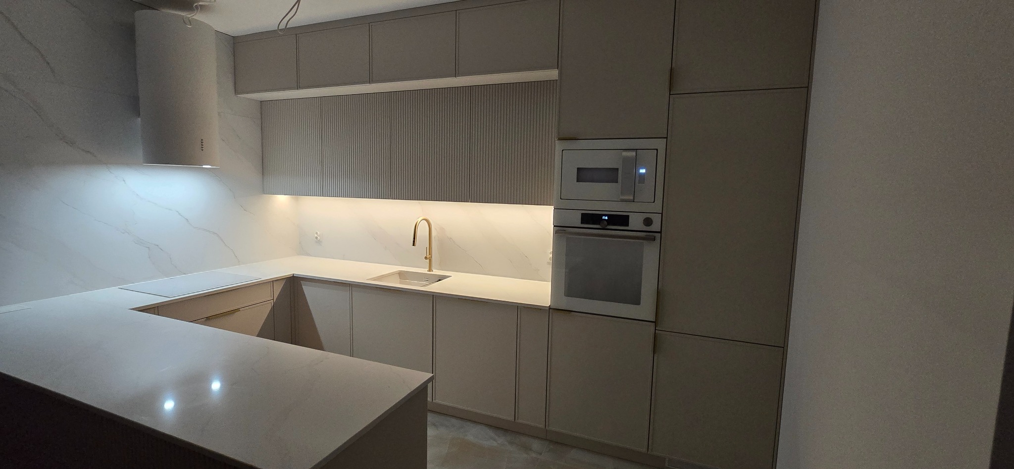 Nowoczesna kuchnia w odcieniach beżu z marmurowym backsplashiem, złotą baterią i oświetleniem LED pod szafkami górnymi.