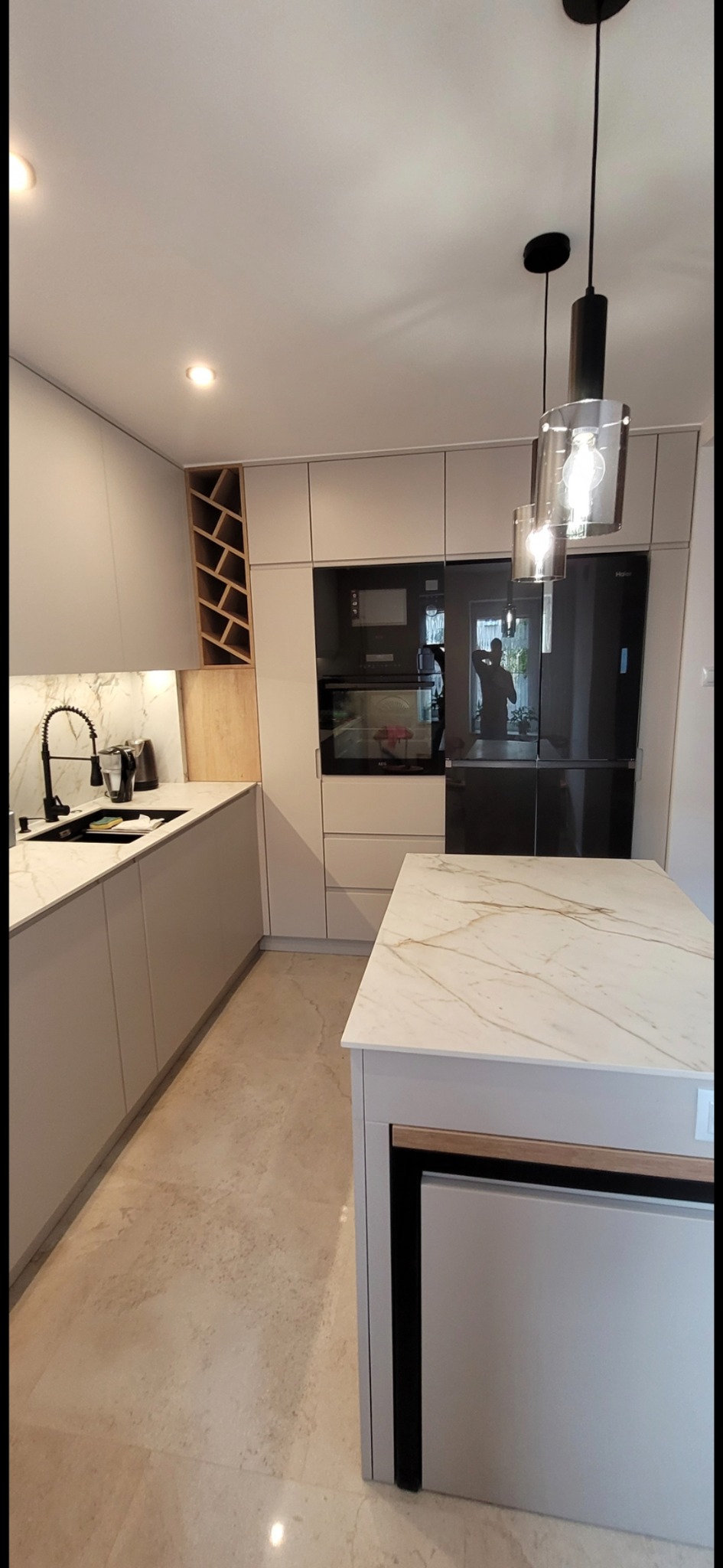 Nowoczesna kuchnia z szarymi frontami, marmurowym blatem i wyspą, oświetlona wiszącymi lampami z przezroczystego szkła, z wbudowanym regałem na wino.