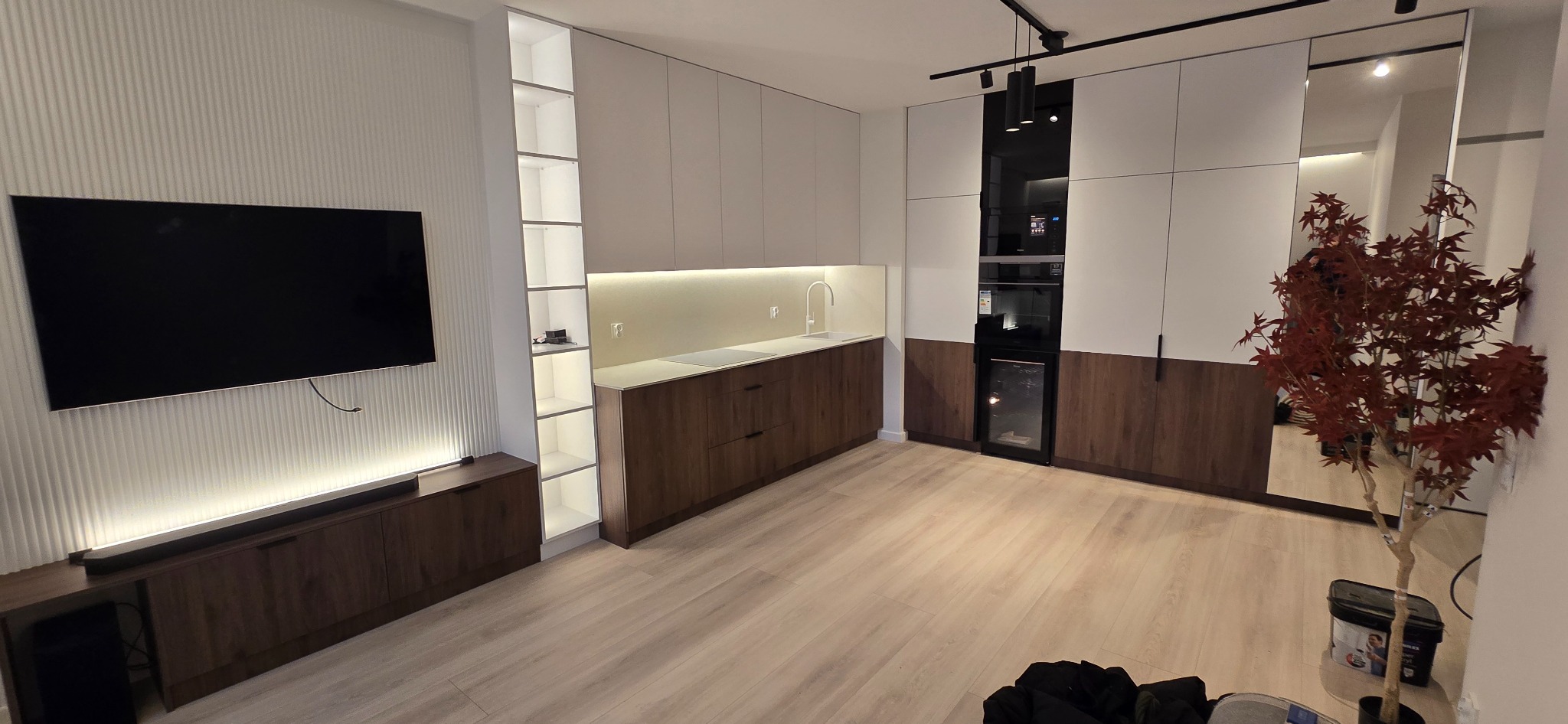 Nowoczesna kuchnia z białymi górnymi szafkami i drewnianymi dolnymi, zintegrowany piekarnik i chłodziarka do wina, oświetlenie LED podszafkowe, minimalistyczny zlew i bateria, oraz dekoracyjny klon...