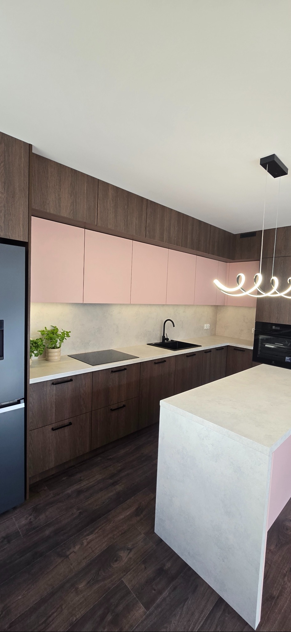 Nowoczesna kuchnia z wyspą: fronty w odcieniach różu i drewna, czarny zlew i płyta, minimalistyczna lampa. Roślina w doniczce dodaje naturalnego akcentu.