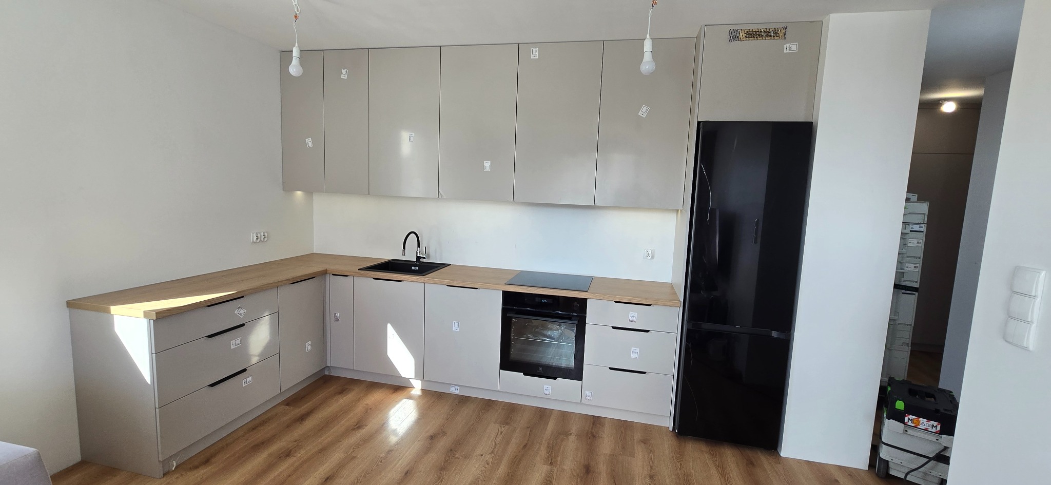 Jasna, nowoczesna kuchnia z drewnianym blatem i czarnymi akcentami: zlew, płyta indukcyjna i lodówka. Widoczne zabezpieczenia na frontach szafek.