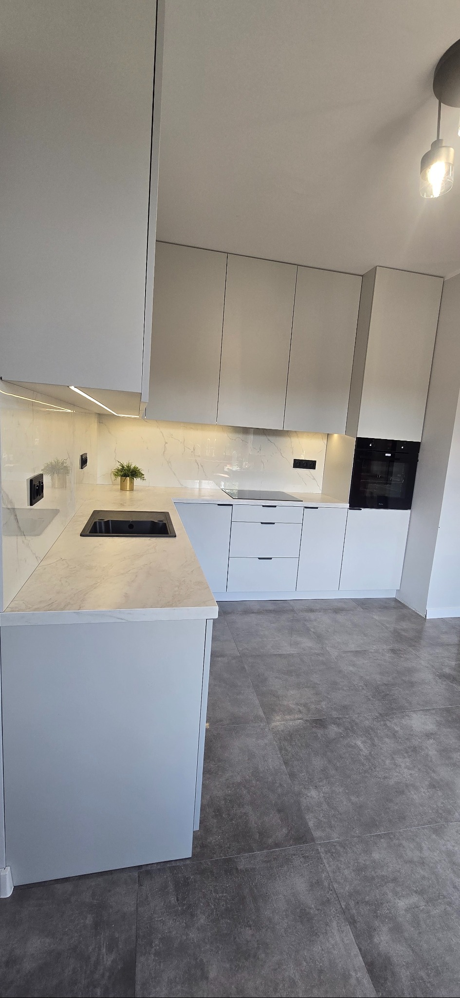 Jasna kuchnia z marmurowym blatem i złotą doniczką z rośliną. Minimalistyczny design, oświetlenie LED pod szafkami, ciemna podłoga. Ujęcie z perspektywy podłogi.