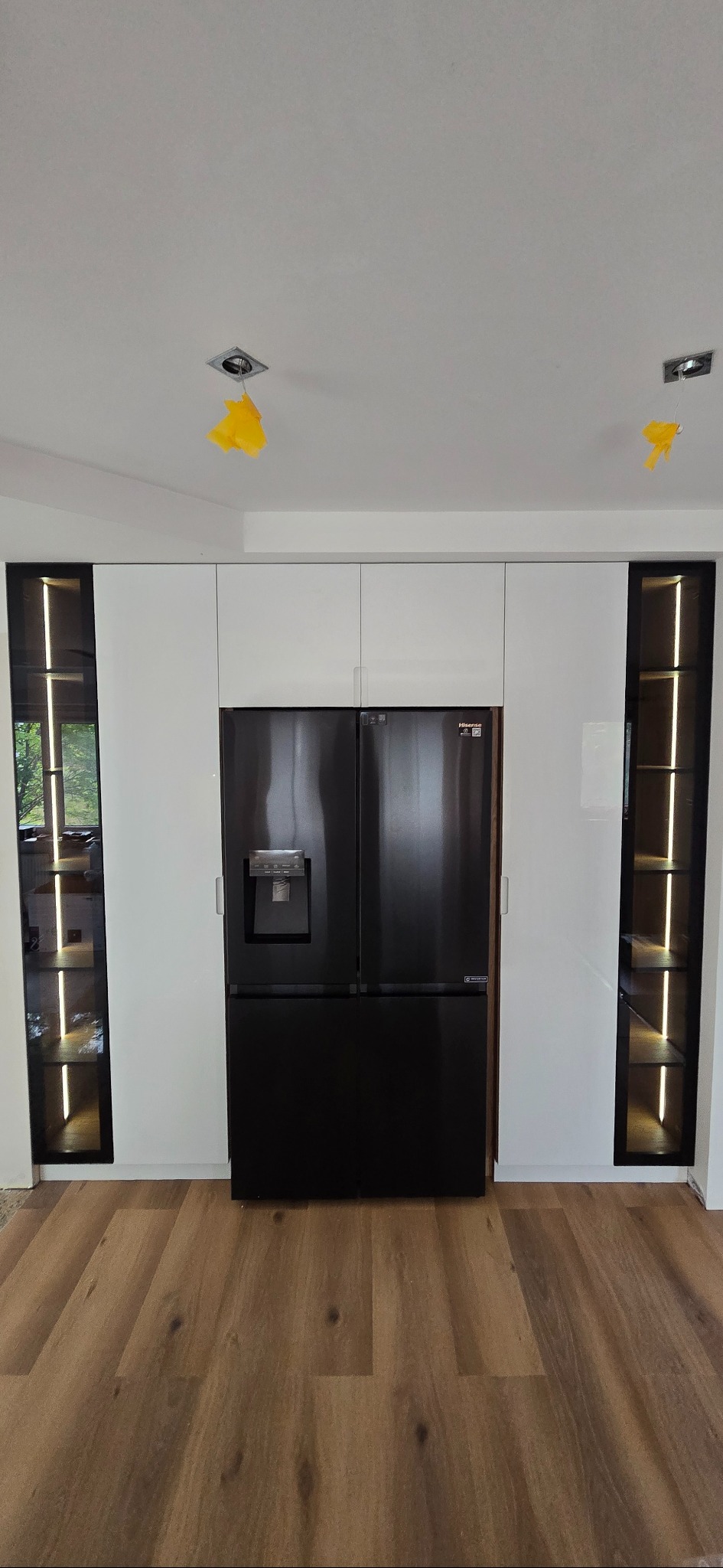 Białe szafki kuchenne z czarną lodówką i podświetlanymi półkami po bokach. Widoczne zabezpieczenia na lampach sufitowych. Parkiet na podłodze.