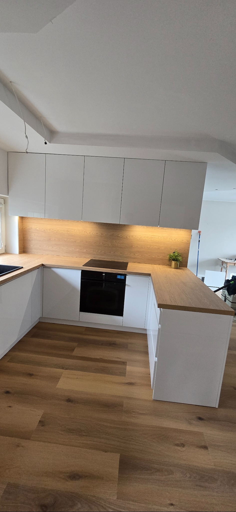 Nowoczesna kuchnia na wymiar z białymi, lakierowanymi frontami i drewnianym blatem. Oświetlenie LED podkreśla ciepło drewna. Widoczna płyta indukcyjna i piekarnik.