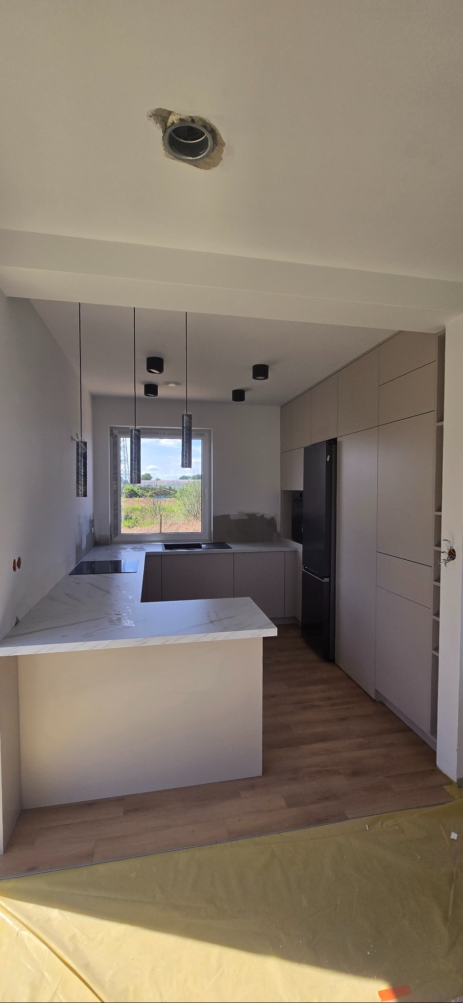 Jasna kuchnia z wyspą i marmurowym blatem w surowym stanie. Widoczne niedokończone oświetlenie sufitowe i zabezpieczona podłoga. Okno z widokiem na zieleń.