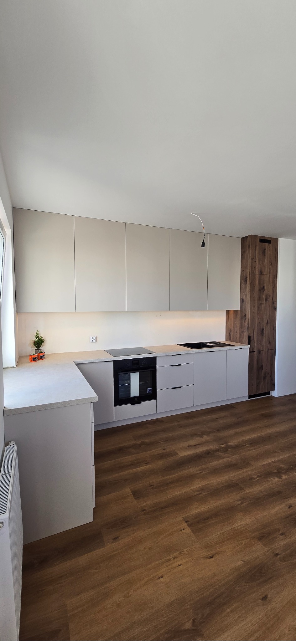 Jasna kuchnia z drewnianym akcentem: minimalistyczne szafki wiszące z podświetleniem LED, zabudowany piekarnik i płyta, drewniana szafa cargo, brązowa podłoga.