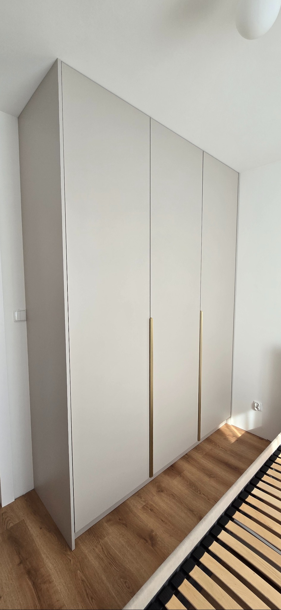 Jasna, minimalistyczna szafa z czterema drzwiami i długimi, złotymi uchwytami, dopasowana do wnętrza sypialni z drewnianą podłogą i widocznym fragmentem łóżka.