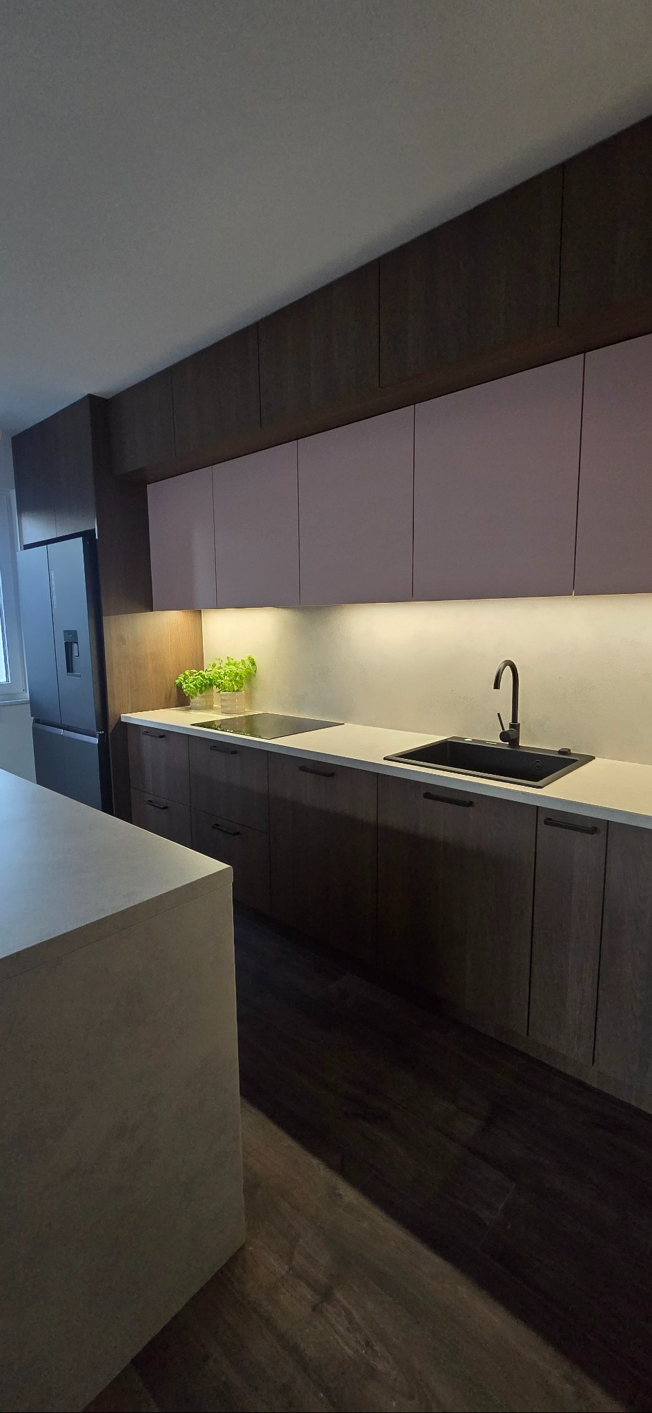 Nowoczesna kuchnia z drewnianymi szafkami, jasnym blatem i subtelnym oświetleniem LED. Uwagę przyciąga minimalistyczna armatura i świeże zioła w doniczce.