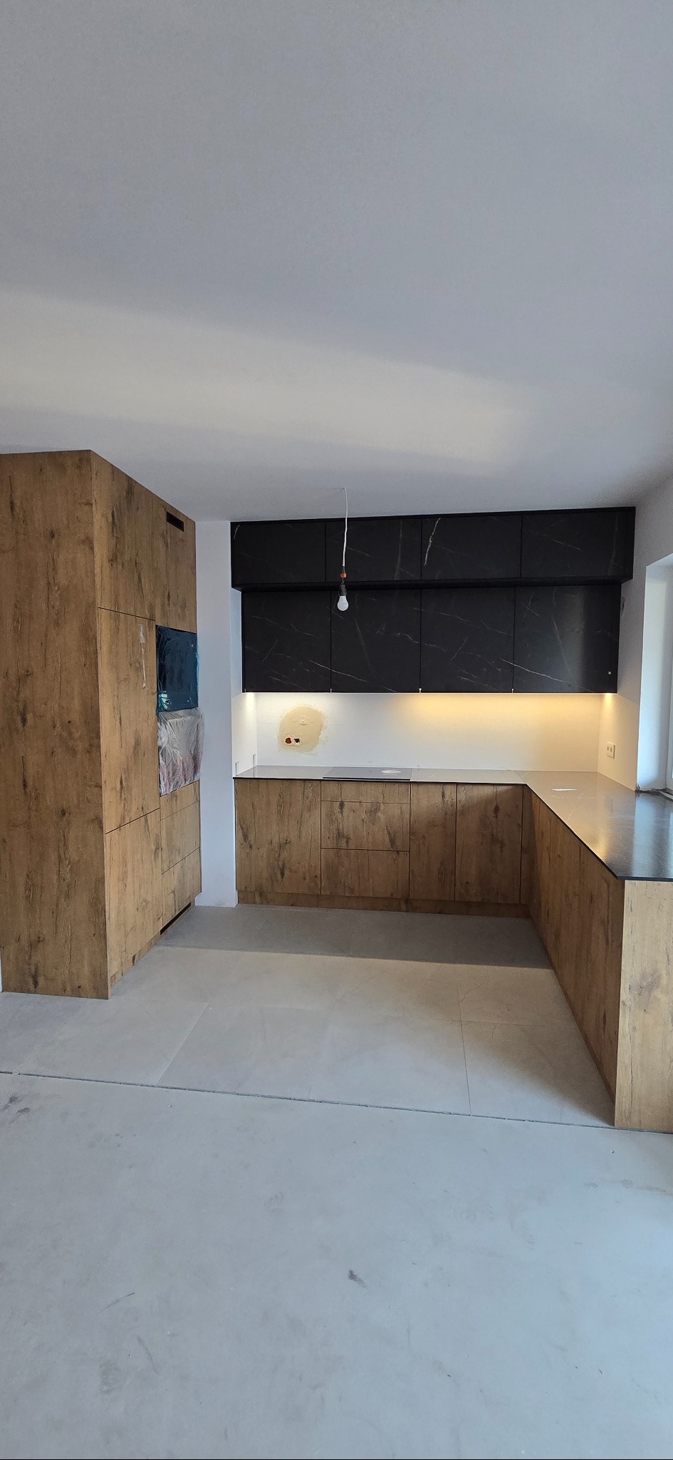 Nowoczesna kuchnia z drewnianymi frontami i czarnymi szafkami wiszącymi z marmurowym wzorem. Widoczne niedokończone prace wykończeniowe.