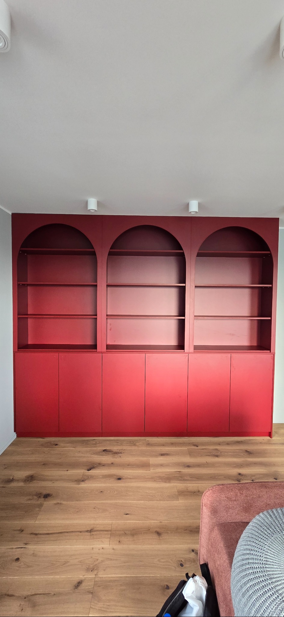 Czerwona zabudowa meblowa z trzema przeszklonymi, łukowatymi wnękami i szafkami na dole, na tle drewnianej podłogi. Minimalistyczny design, nowoczesne oświetlenie.