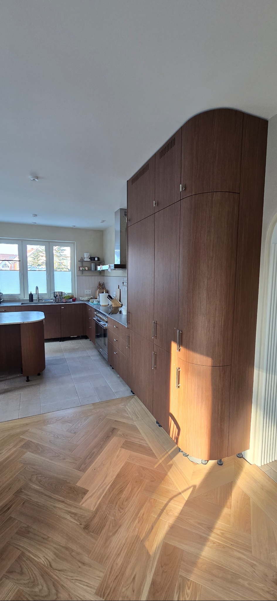 Kuchnia na wymiar z drewnianymi frontami i zaokrąglonym narożnikiem szafy, parkiet w jodełkę, jasne wnętrze z oknem. Nowoczesny design.