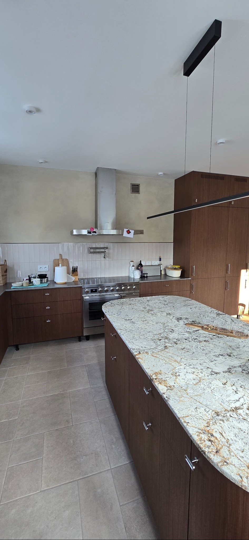 Nowoczesna kuchnia z wyspą o blacie z granitu. Brązowe szafki z poziomym usłojeniem, stalowy okap i armatura. Minimalistyczna lampa sufitowa.