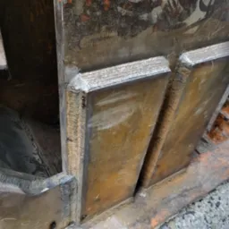 Metalowa konstrukcja z widocznymi spawami, surowe wykończenie, widoczne ślady korozji i zabrudzenia, szara nawierzchnia w tle.