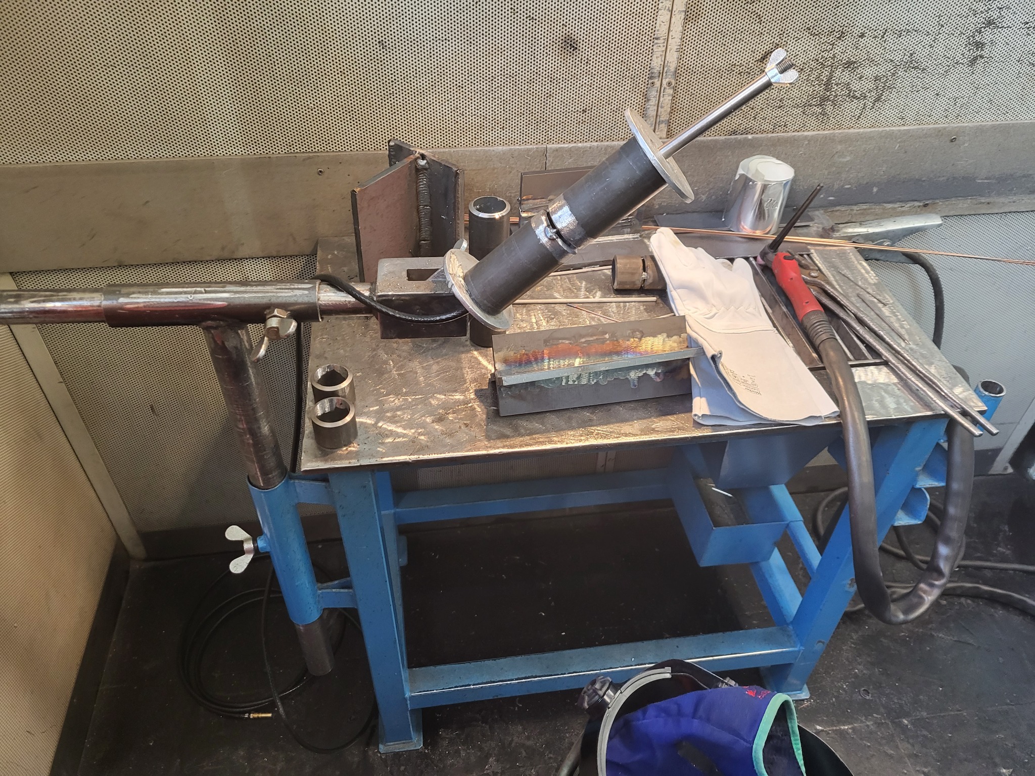 Stanowisko spawalnicze z uchwytem do spawania rur, rękawicami ochronnymi i narzędziami na niebieskim stole warsztatowym, widok z góry.