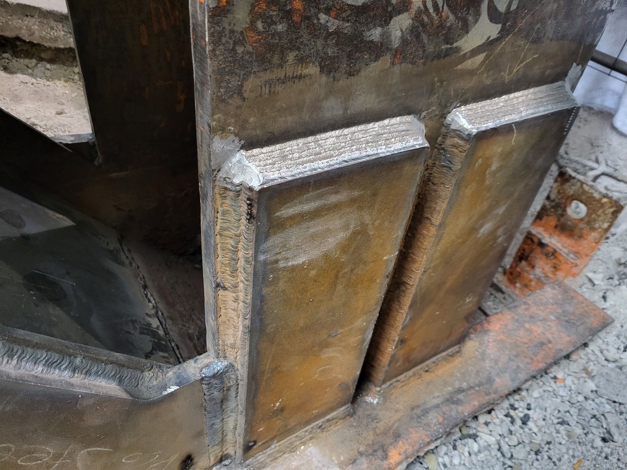 Metalowa konstrukcja z widocznymi spawami, surowe wykończenie, widoczne ślady korozji i zabrudzenia, szara nawierzchnia w tle.