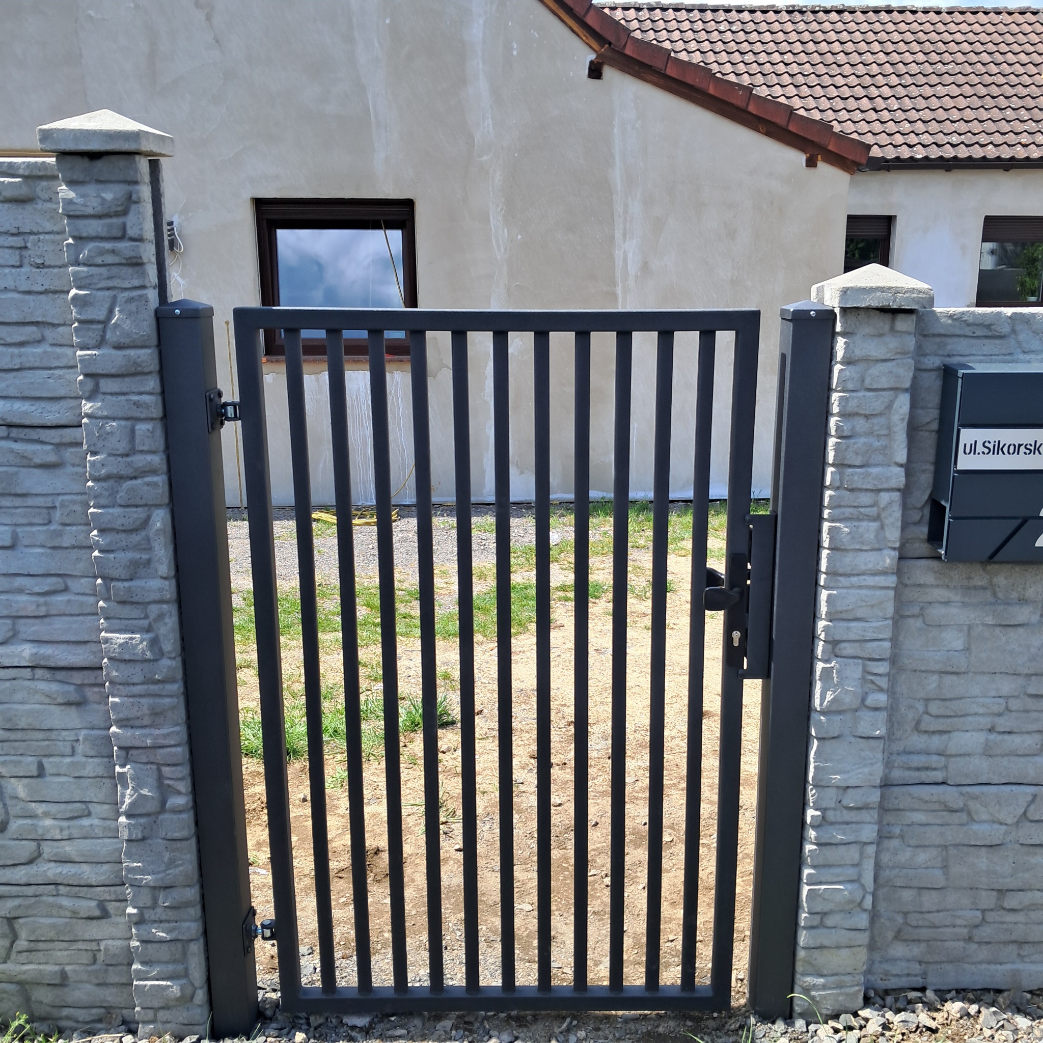 Metalowa furtka z pionowymi prętami, zamontowana między betonowymi słupkami. W tle dom z brązowym dachem i oknem. Adres na skrzynce pocztowej.