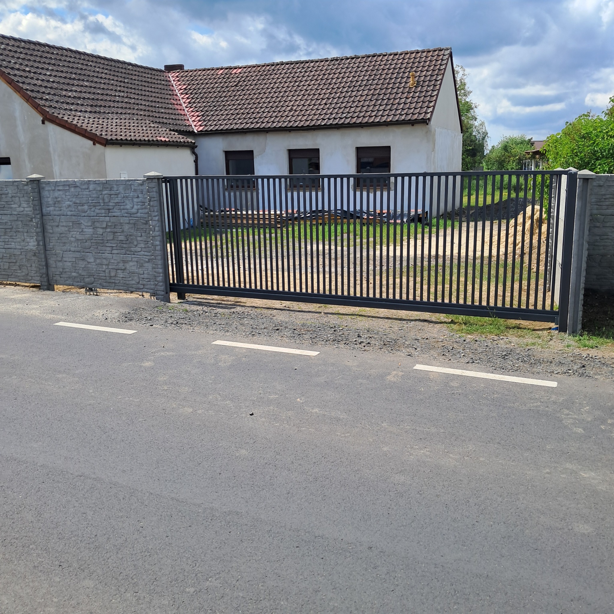 Nowoczesna, przesuwna, metalowa brama wjazdowa z pionowymi szczebelkami, zamontowana przy posesji z betonowym ogrodzeniem i domem w tle. Widok z ulicy.