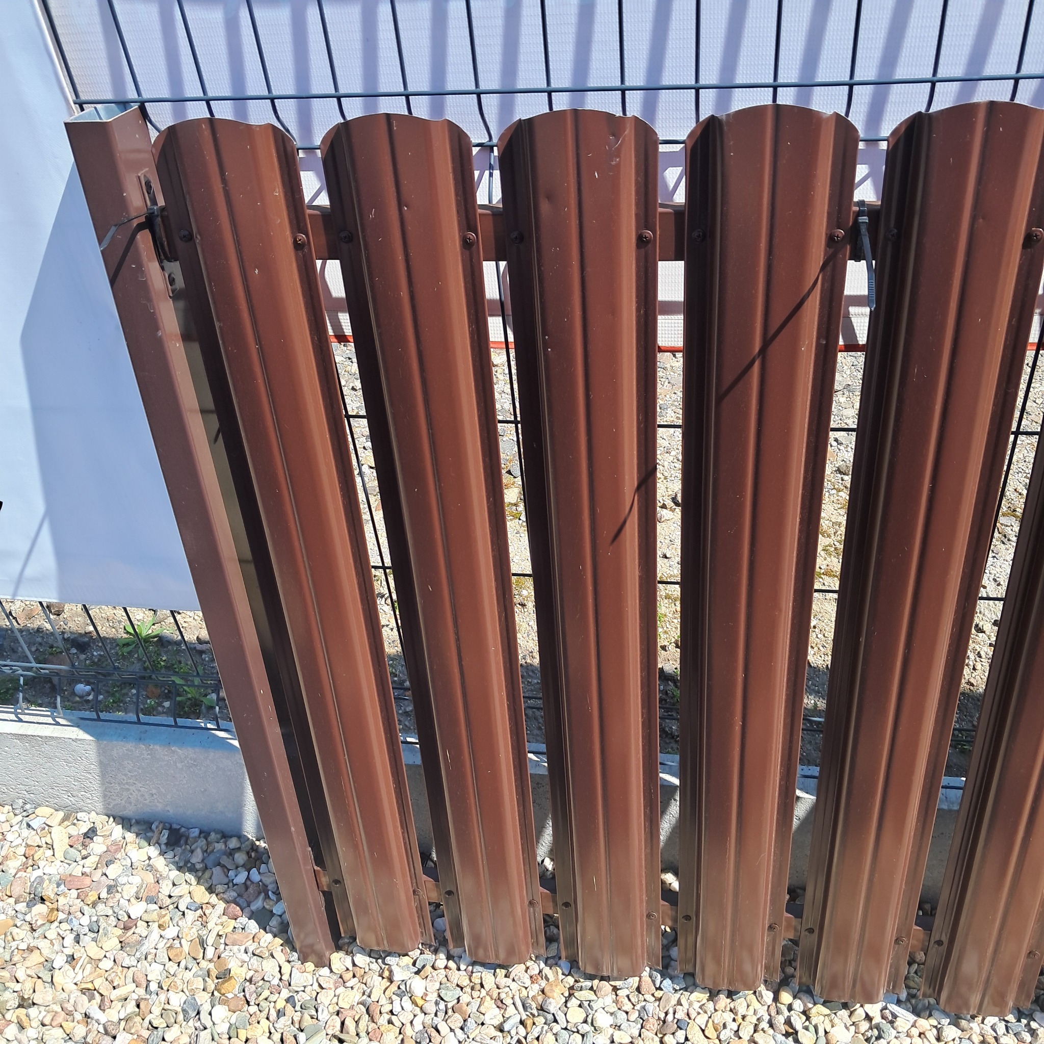 Brązowe, metalowe przęsła ogrodzeniowe z widocznymi śrubami mocującymi, ustawione na żwirowym podłożu, tworzące fragment ogrodzenia panelowego.