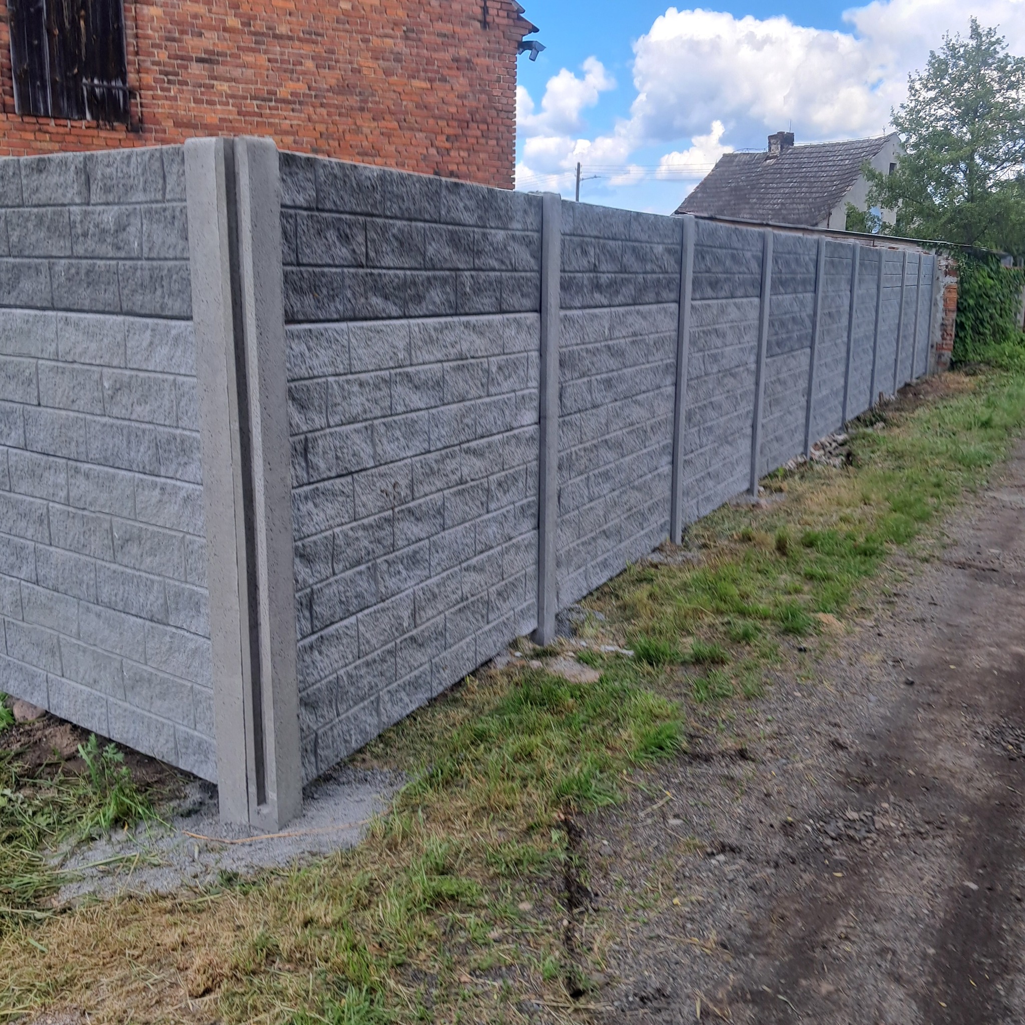 Nowoczesne ogrodzenie betonowe imitujące cegłę, z szarymi słupkami, otoczone zielenią i szarą nawierzchnią. Widok na dom w tle.