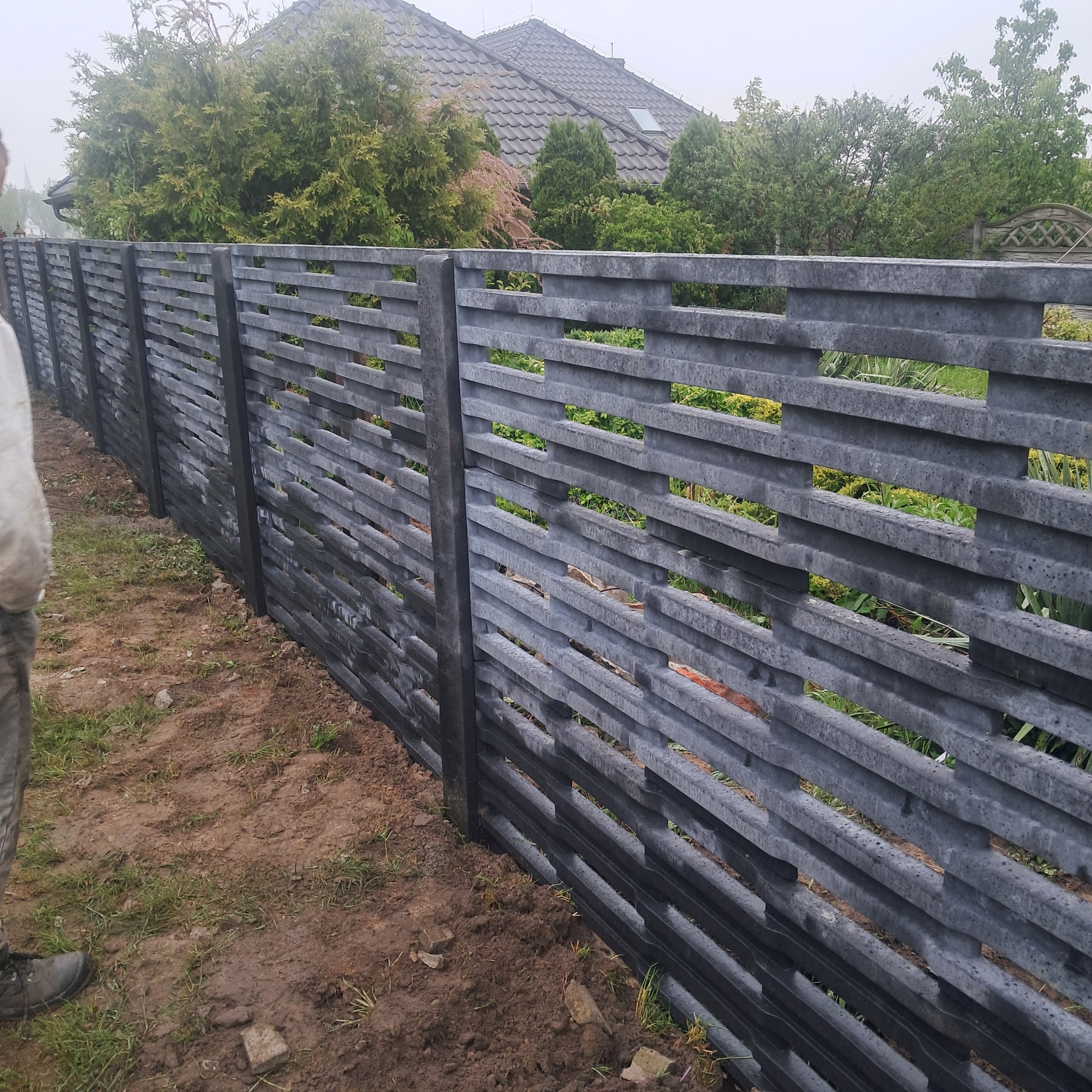 Nowoczesne, szare ogrodzenie betonowe z poziomymi, ażurowymi przęsłami. W tle zieleń ogrodu i dom z szarą dachówką. Widok z bliska na nowo postawione ogrodzenie.