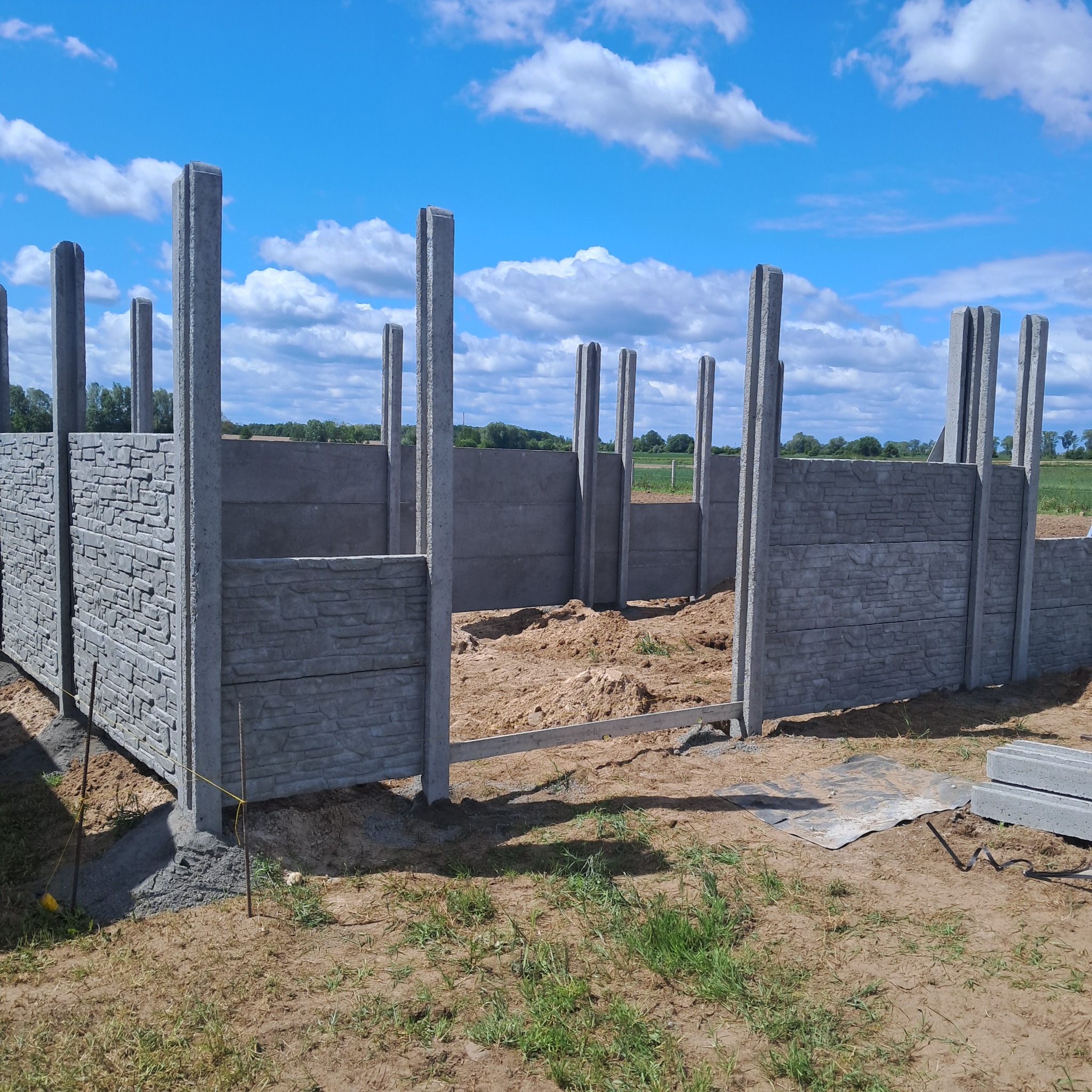 Montaż ogrodzenia betonowego z imitacją kamienia, widoczne słupki i panele w trakcie instalacji na tle błękitnego nieba z chmurami.