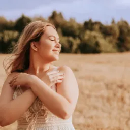 Uśmiechnięta kobieta w białej sukience, obejmująca się ramionami na tle pola i drzew, z delikatnym oświetleniem i miękkimi cieniami.