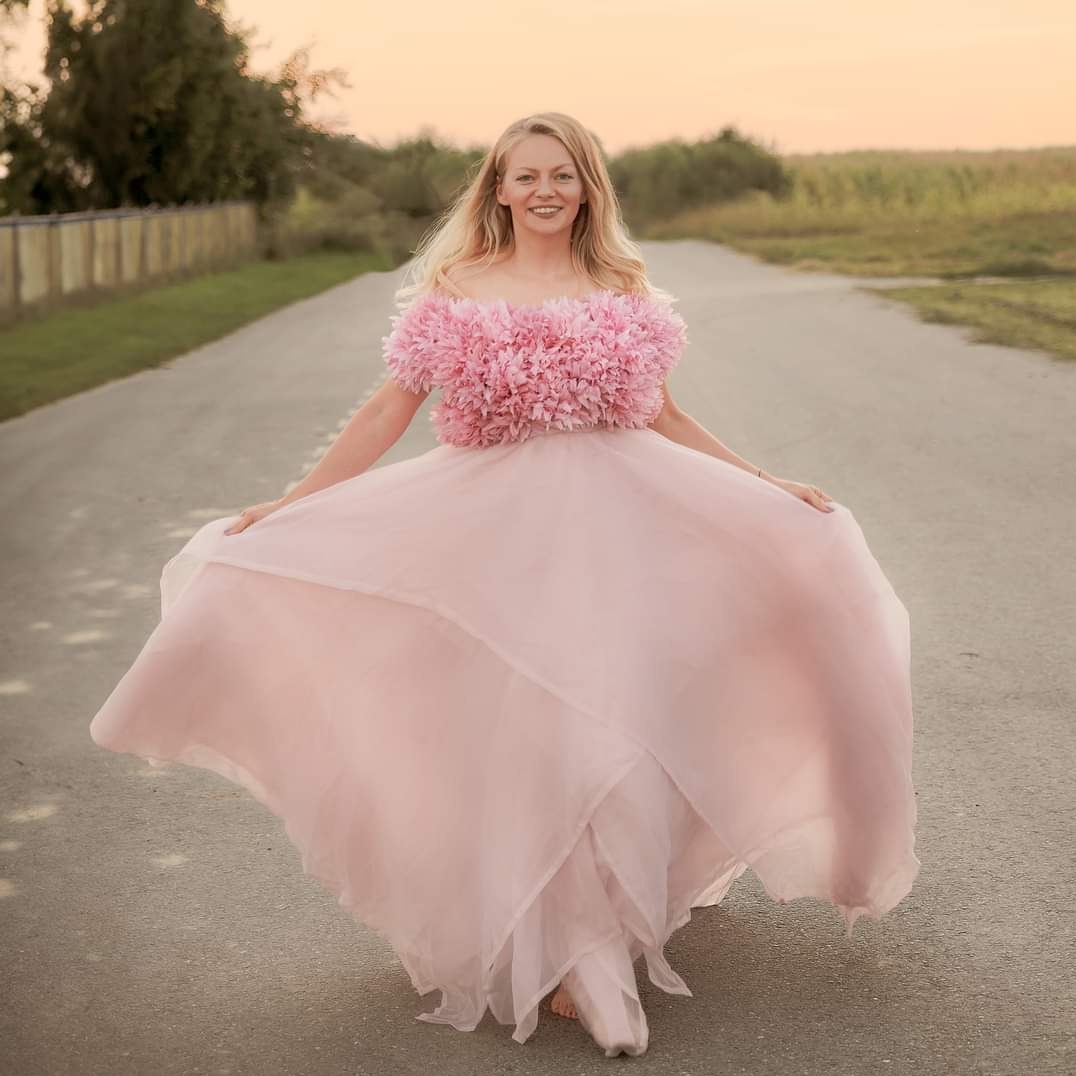 Uśmiechnięta blondynka w zwiewnej, różowej sukni z kwiatowym topem, spacerująca boso po asfaltowej drodze w wiejskim krajobrazie o zachodzie słońca.