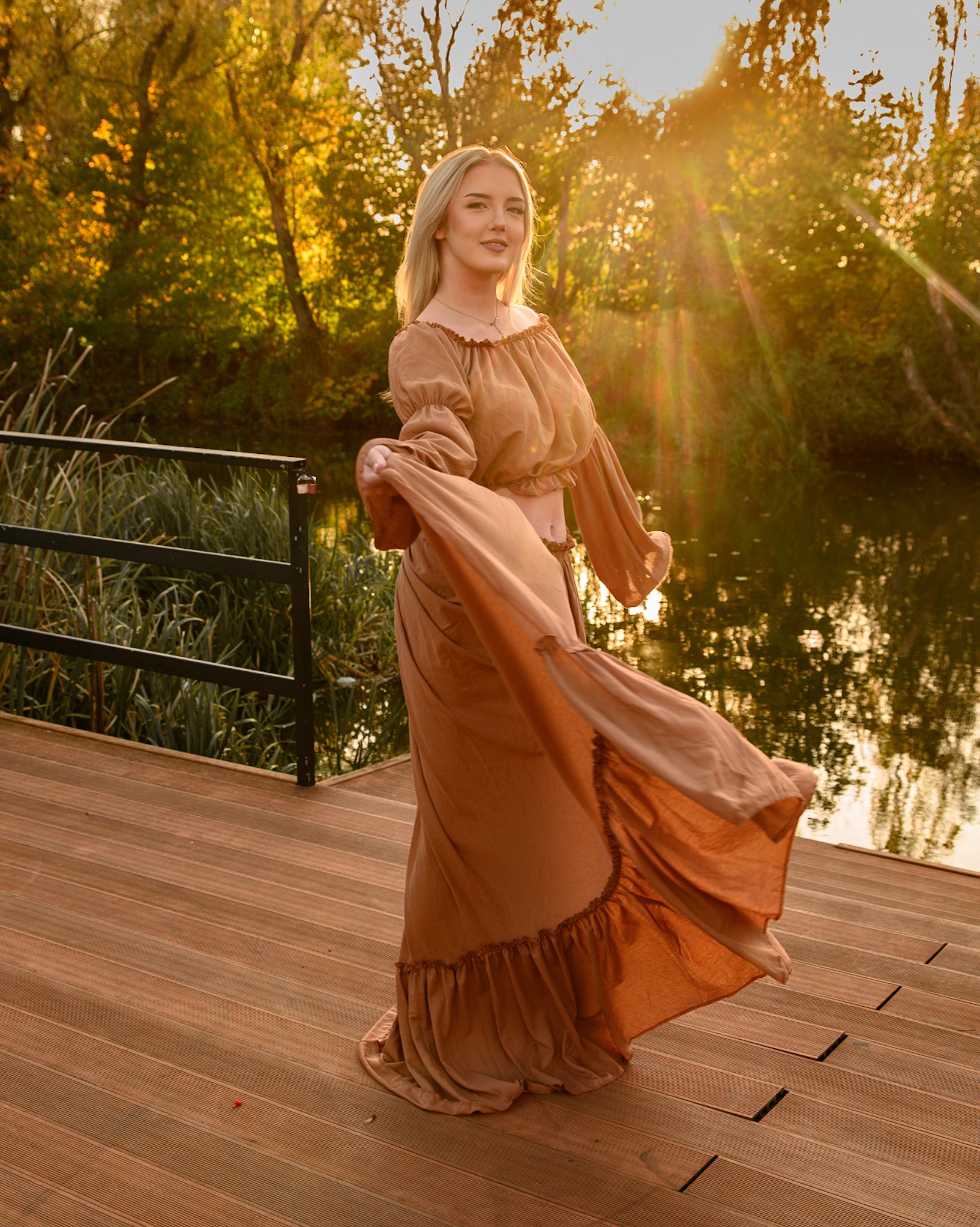 Kobieta w zwiewnej, brązowej sukience pozuje na drewnianym pomoście nad wodą, z promieniami zachodzącego słońca w tle.