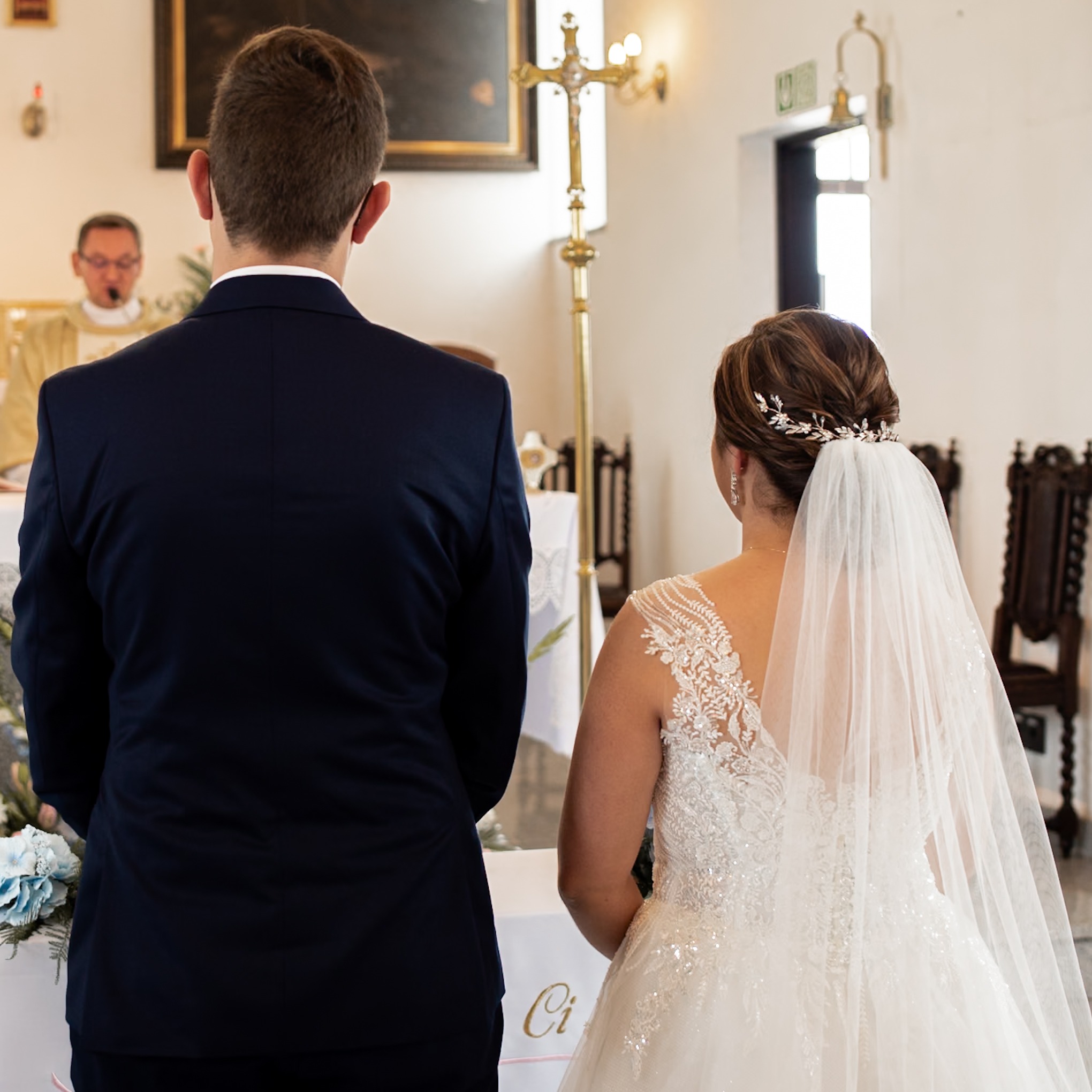 Para młoda podczas ceremonii ślubnej w kościele, widok z tyłu, panna młoda w sukni z koronkowymi aplikacjami i welonem, pan młody w granatowym garniturze, w tle ksiądz przy ołtarzu.