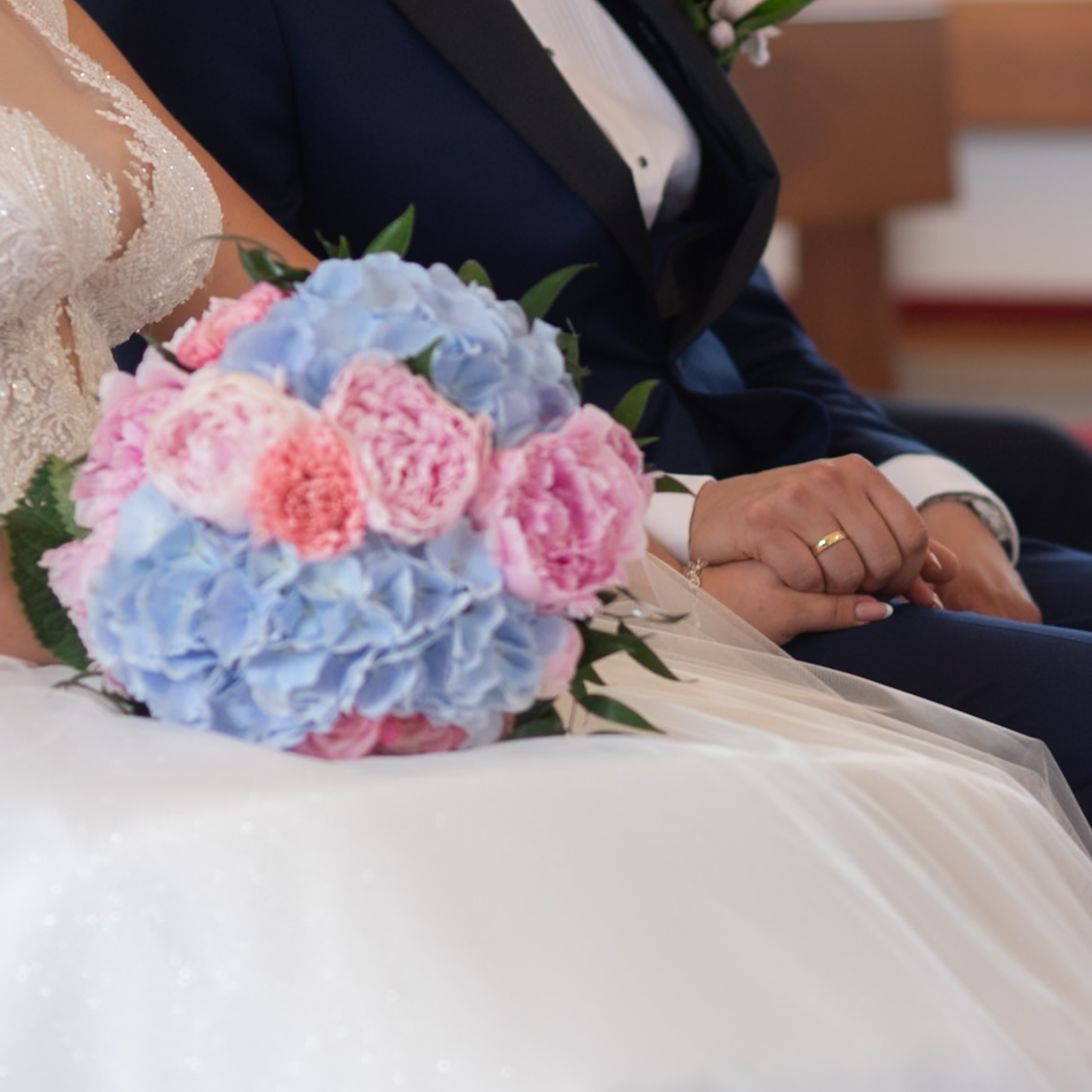 Para młoda trzymająca się za ręce w kościele, widoczny bukiet z niebieskimi i różowymi kwiatami oraz złota obrączka na palcu.