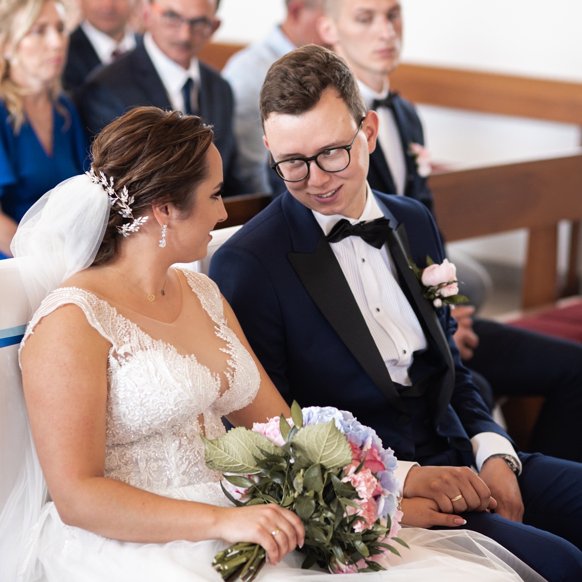 Uśmiechnięta para młoda siedzi obok siebie w kościele, panna młoda trzyma bukiet z różowych i niebieskich kwiatów, pan młody w granatowym smokingu z czarną muchą i różową butonierką, w tle goście...