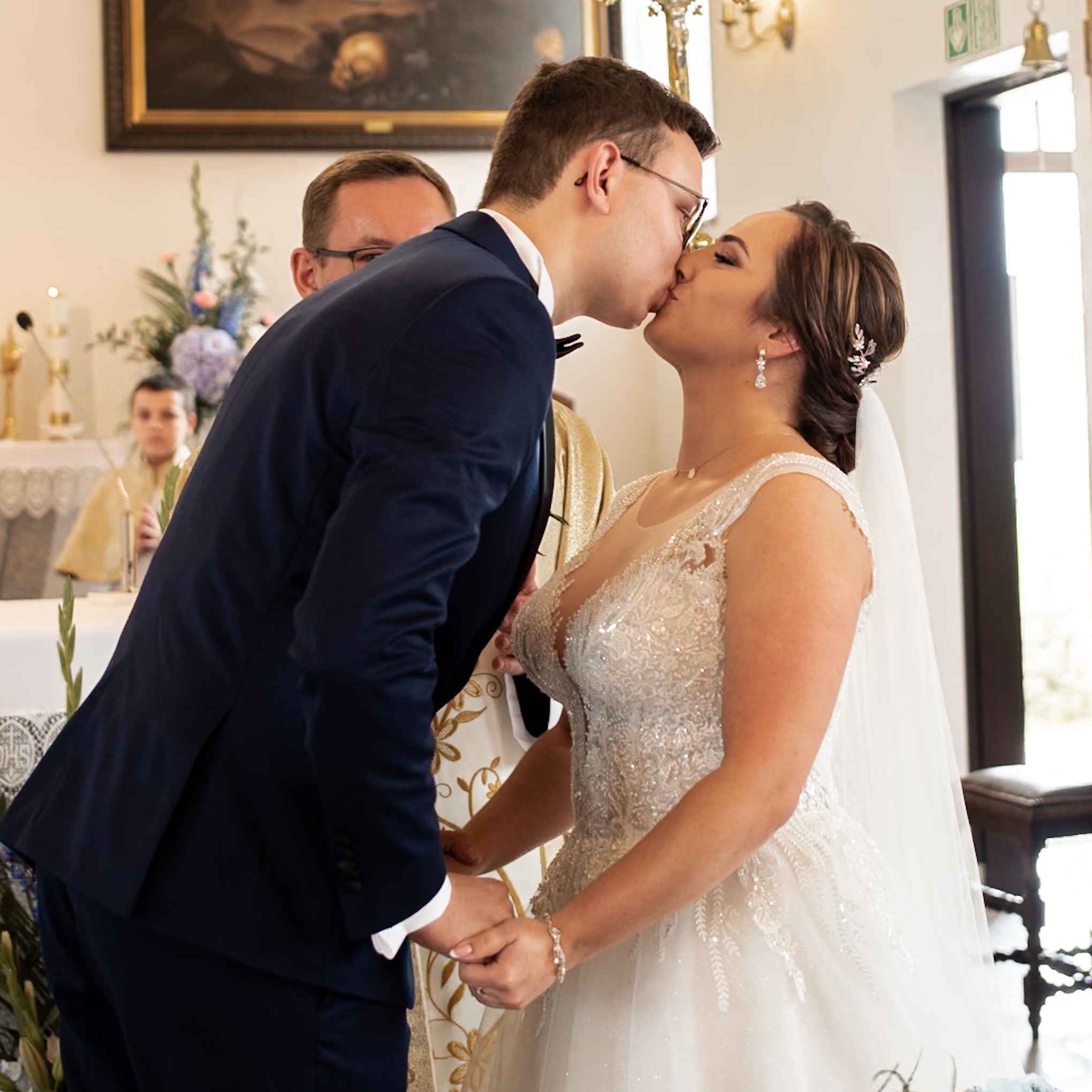 Pocałunek młodej pary podczas ceremonii ślubnej w kościele, w tle ksiądz i ministrant, panna młoda w bogato zdobionej sukni z koronką i welonem, pan młody w granatowym garniturze z muchą.