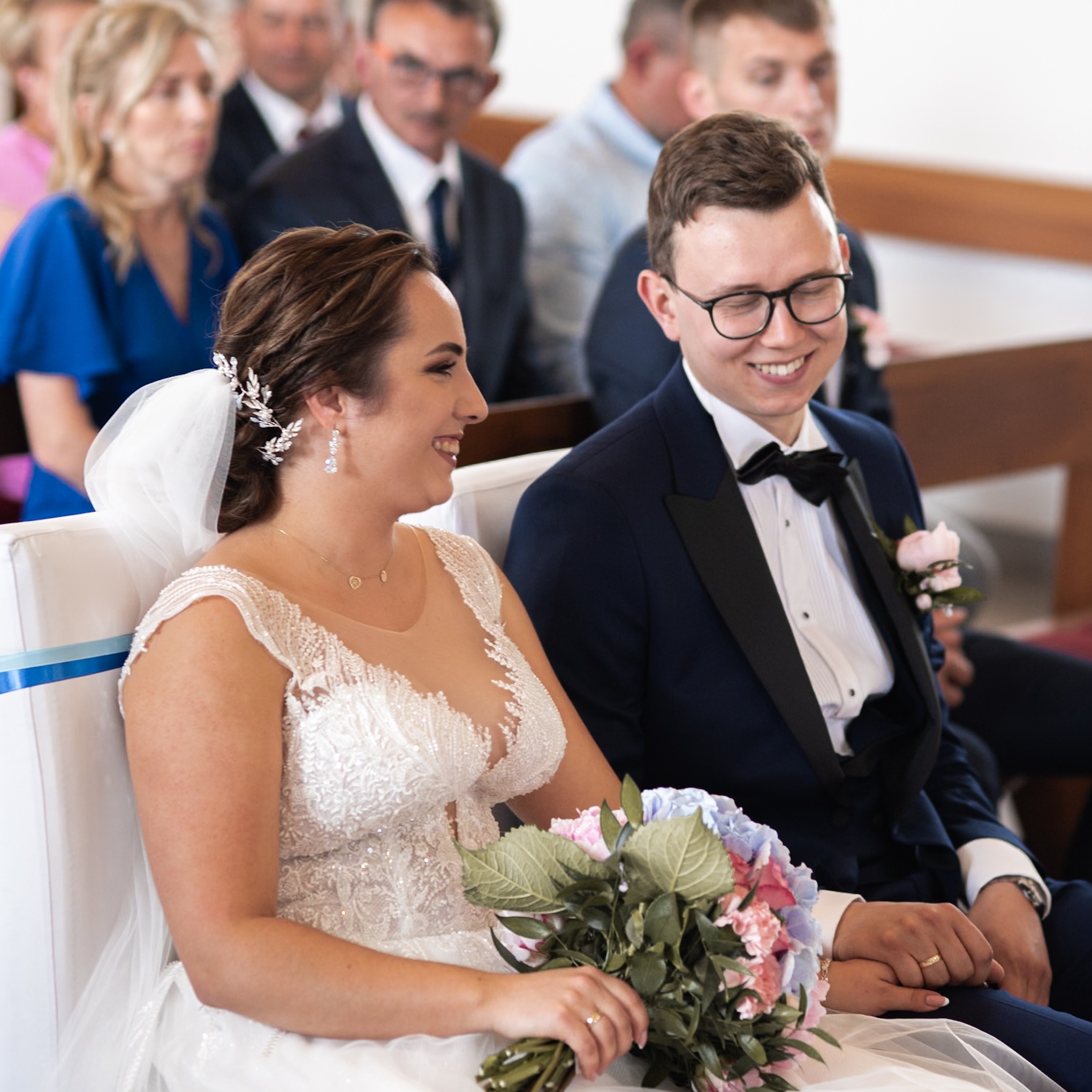 Uśmiechnięta para młoda siedzi w kościele podczas ceremonii, panna młoda trzyma bukiet różowych i niebieskich kwiatów, a pan młody ma na sobie okulary.
