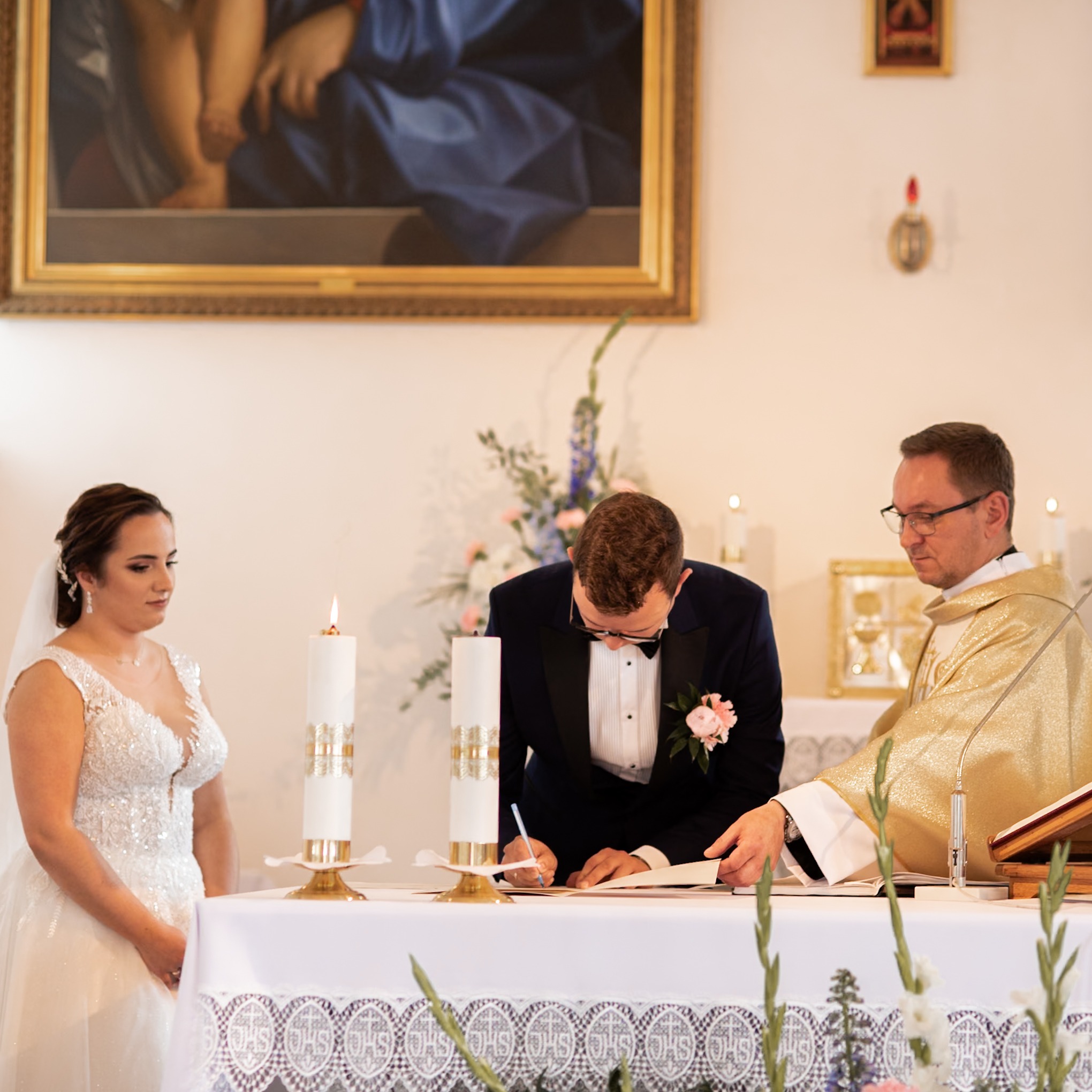 Para młoda podpisuje dokumenty podczas ceremonii ślubnej w kościele, w tle ksiądz w złotej szacie i obraz religijny