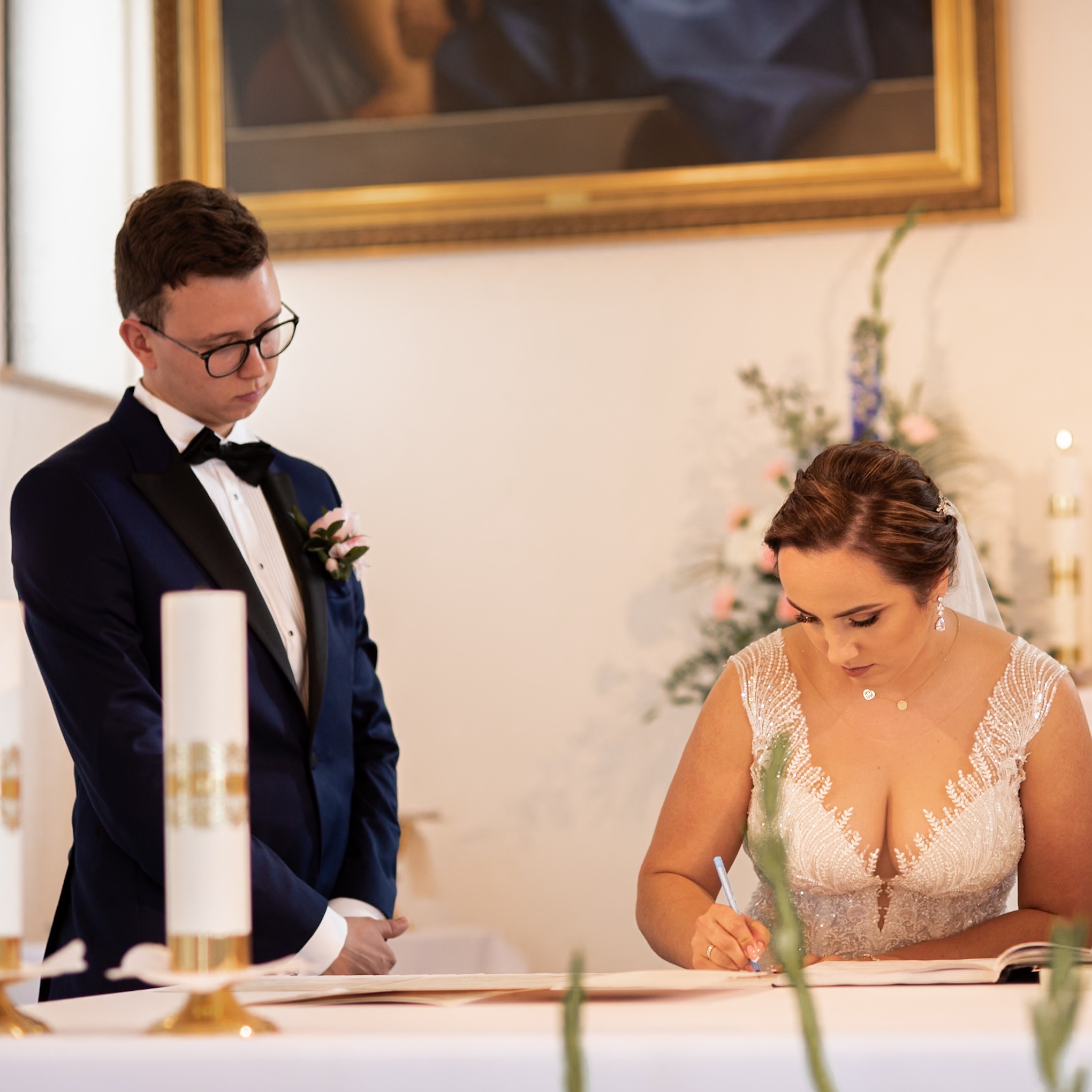 Panna młoda w sukni z koronkowym dekoltem podpisuje dokument, obok stoi pan młody w granatowym smokingu z bukiecikiem, w tle obraz w złotej ramie i kwiatowa dekoracja.