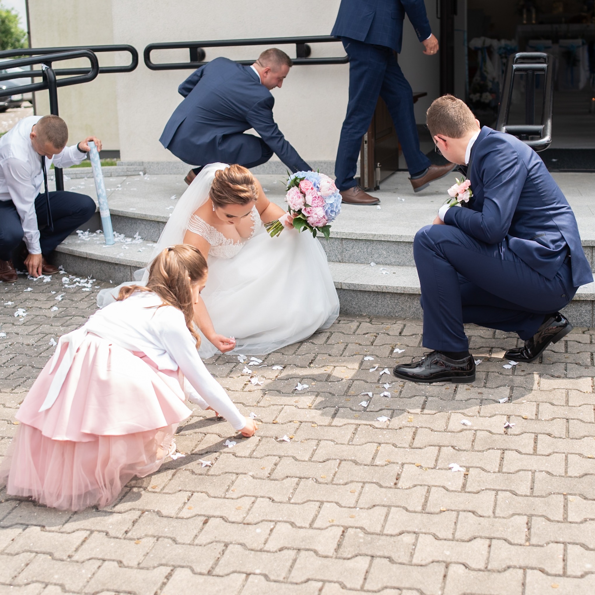 Panna młoda w białej sukni z bukietem różowych i niebieskich kwiatów, klęczy na chodniku przed kościołem zbierając konfetti z pomocą drużby i małej dziewczynki w różowej sukience, podczas gdy inni...