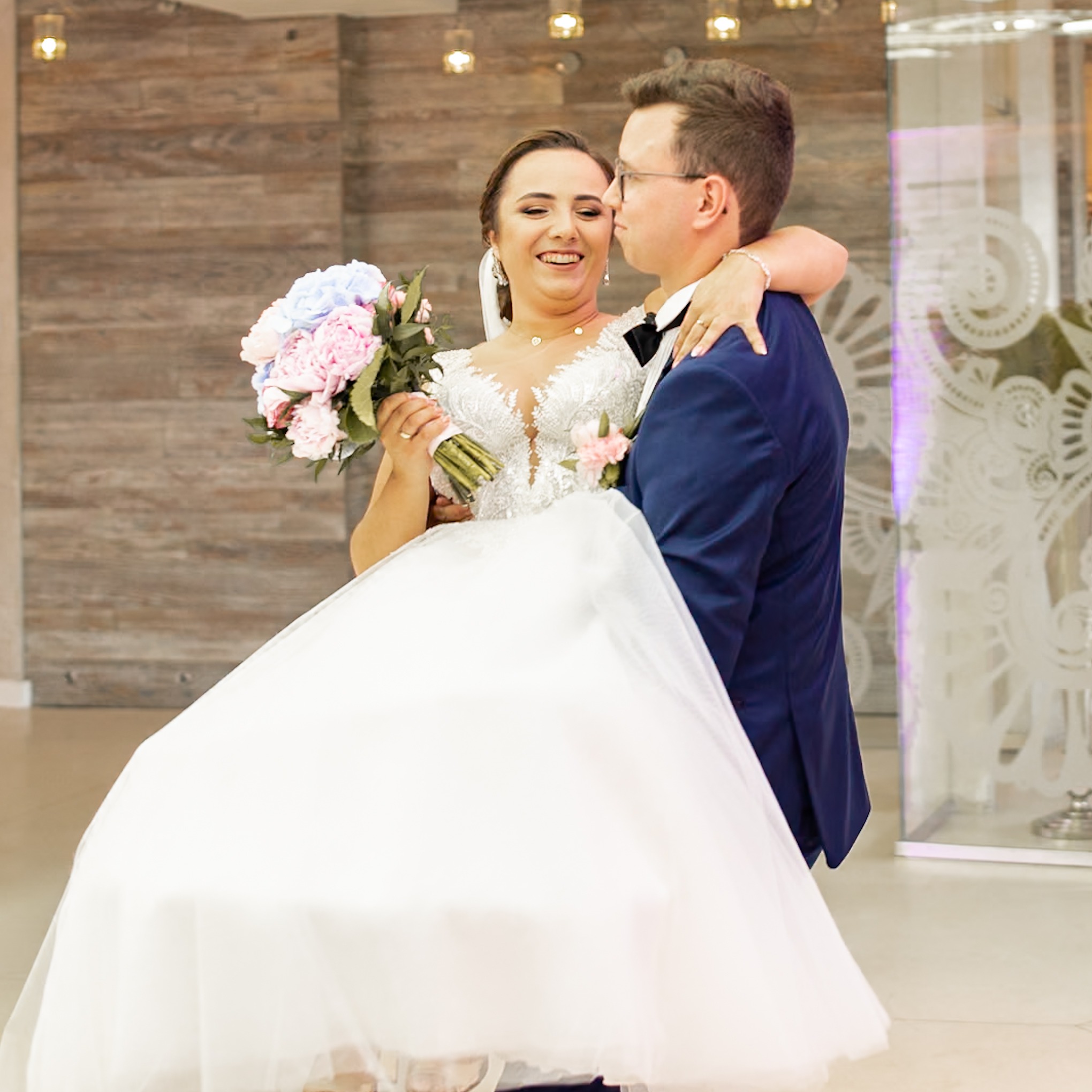 Uśmiechnięta panna młoda w białej sukni z bukietem różowych i niebieskich kwiatów, podnoszona przez pana młodego w granatowym garniturze, który ją całuje w policzek, na tle drewnianej ściany...