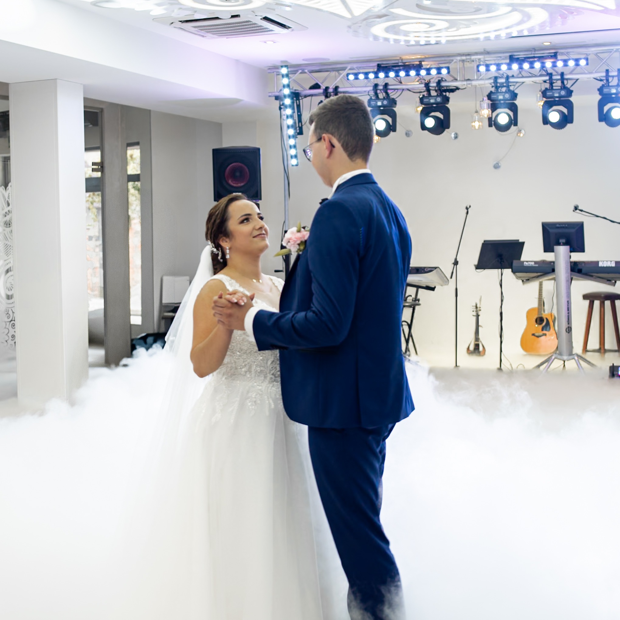 Para młoda w tańcu na tle sztucznej mgły, oświetlenia scenicznego i instrumentów muzycznych.