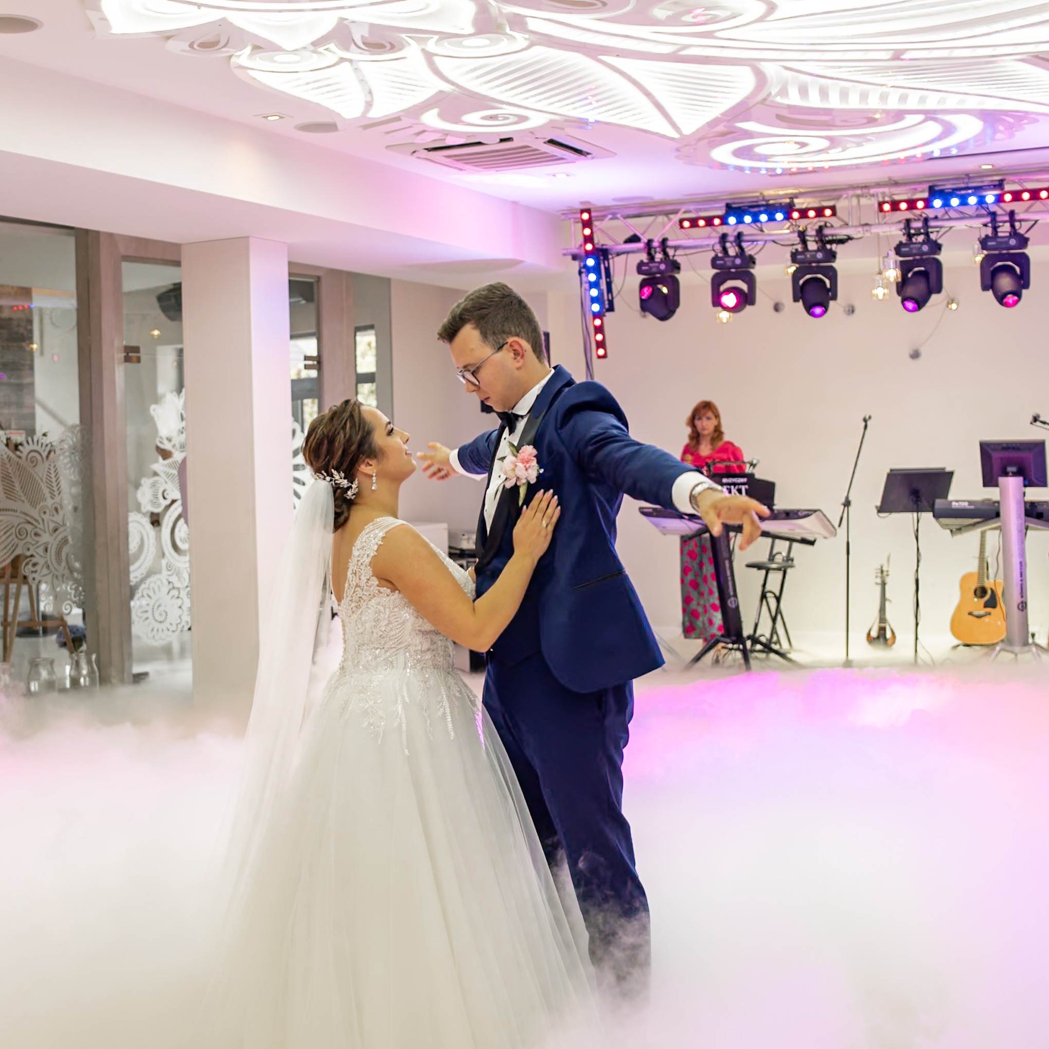 Para młoda w tańcu na sali weselnej, otoczona sztuczną mgłą, w tle zespół muzyczny z instrumentami.