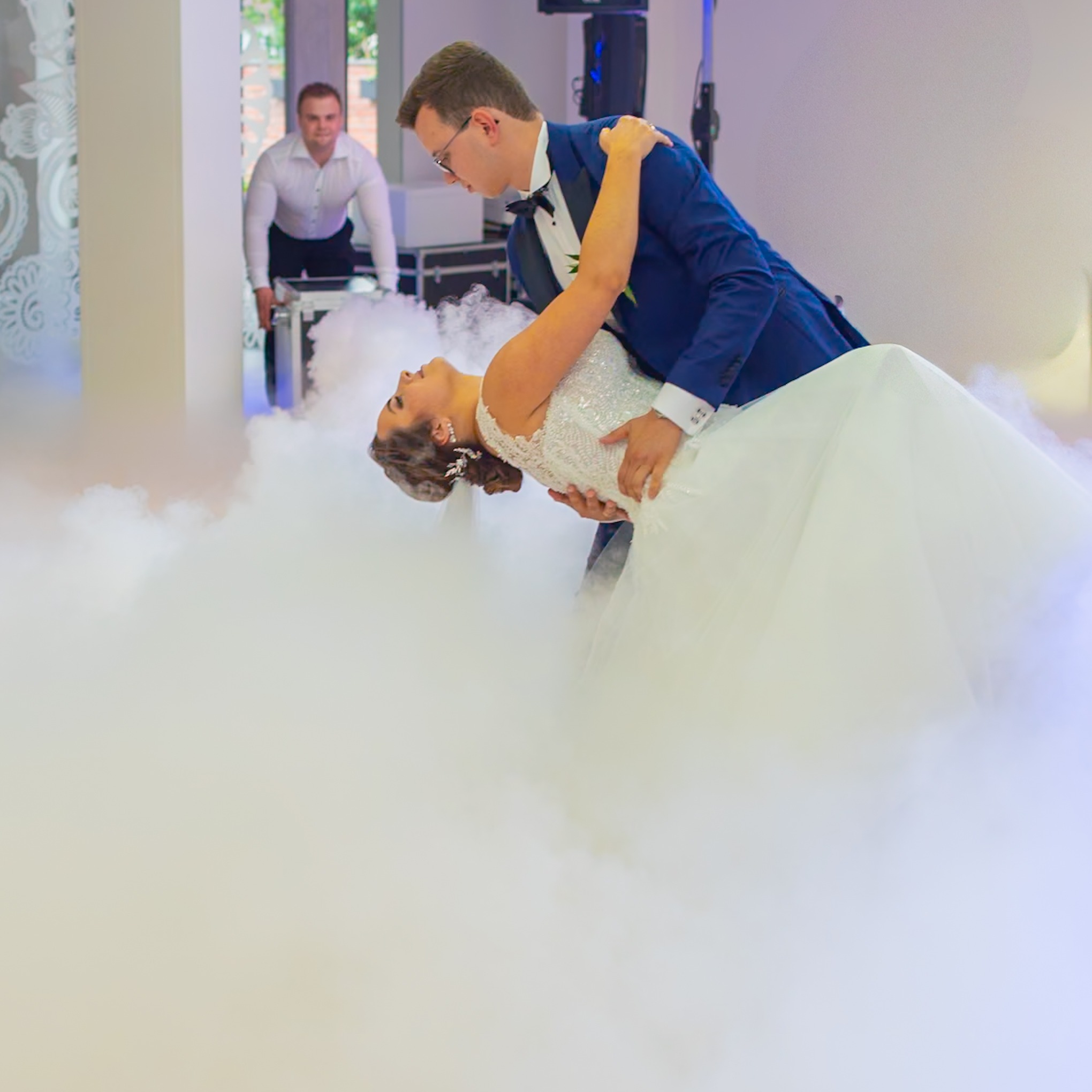 Para młoda w tańcu, mężczyzna w granatowym garniturze podtrzymuje kobietę w białej sukni, unosząc ją w obłokach sztucznego dymu. W tle mężczyzna przywozi sprzęt.